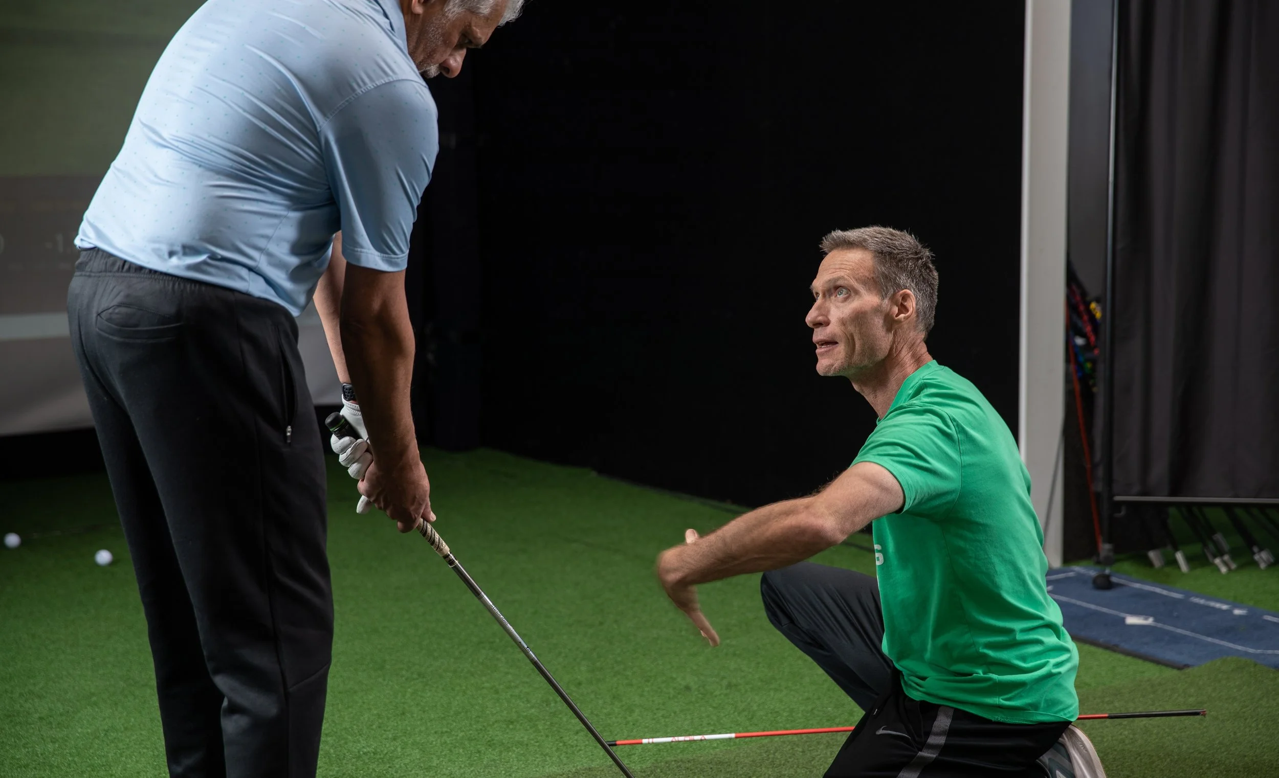 A golf instructor is kneeling on the floor, explaining to a man holding a golf club how to improve his golf swing on an indoor practice mat.