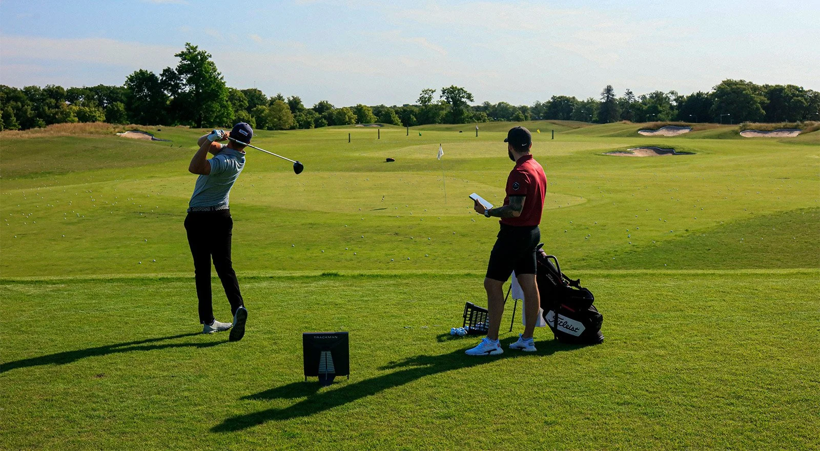 A golfer swinging a club on a golf course while another person watches with a clipboard near a bag of golf clubs.