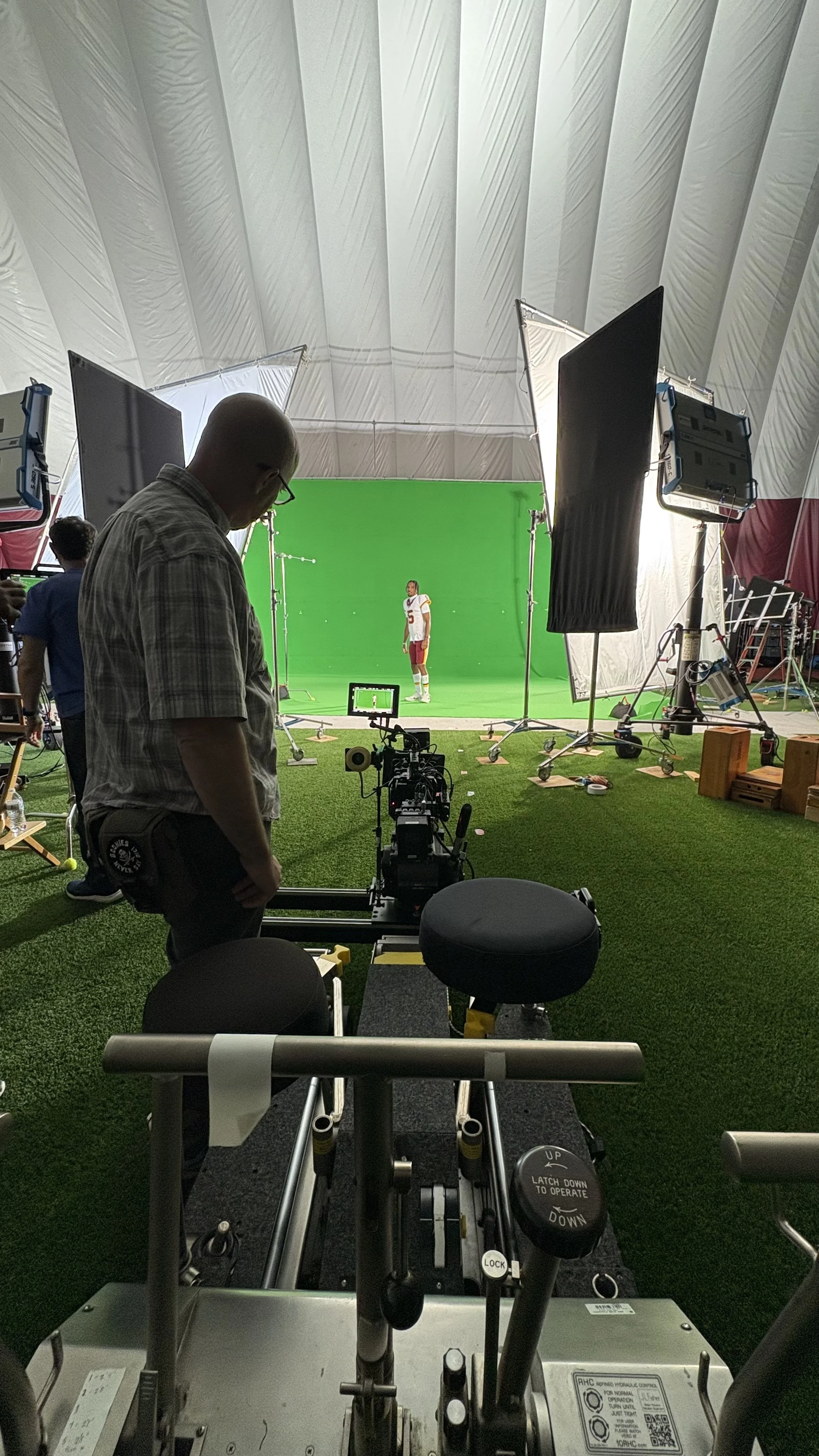 A behind-the-scenes shot of a film or photo shoot setup inside a large tent with a person working in the foreground and a man standing near a camera in front of a green screen with professional lighting equipment surrounding a football player in a je