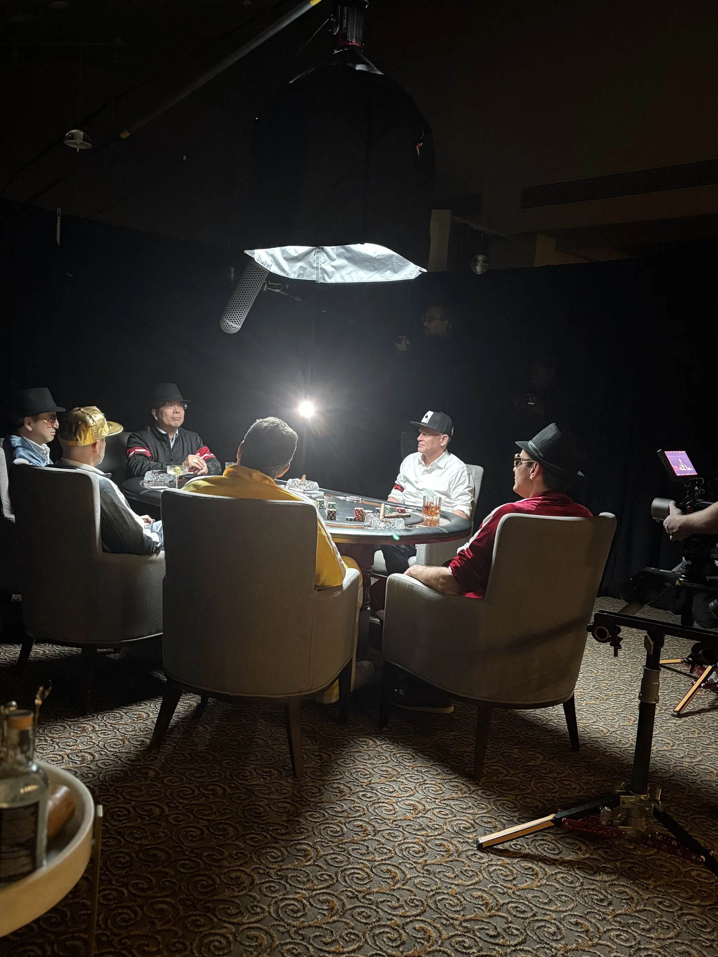 A group of people sitting around a poker table with a large softbox light above them, with camera equipment set up nearby.