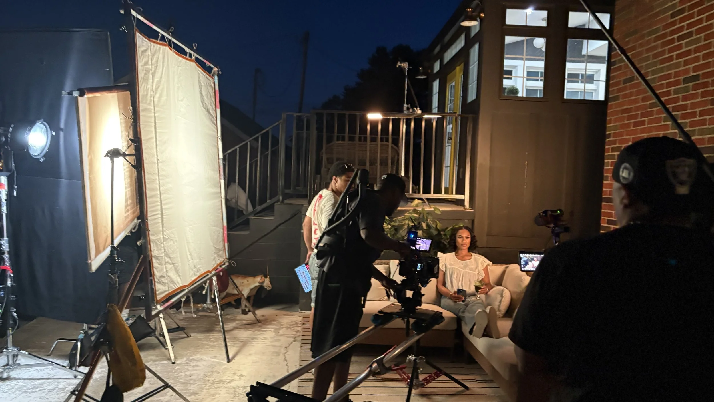 Scene from a film or photo shoot at an outdoor patio at night, with crew members setting up a camera and lighting. A woman in a white outfit sits on a couch holding a glass, while crew members work around her with equipment and monitors. The backgrou