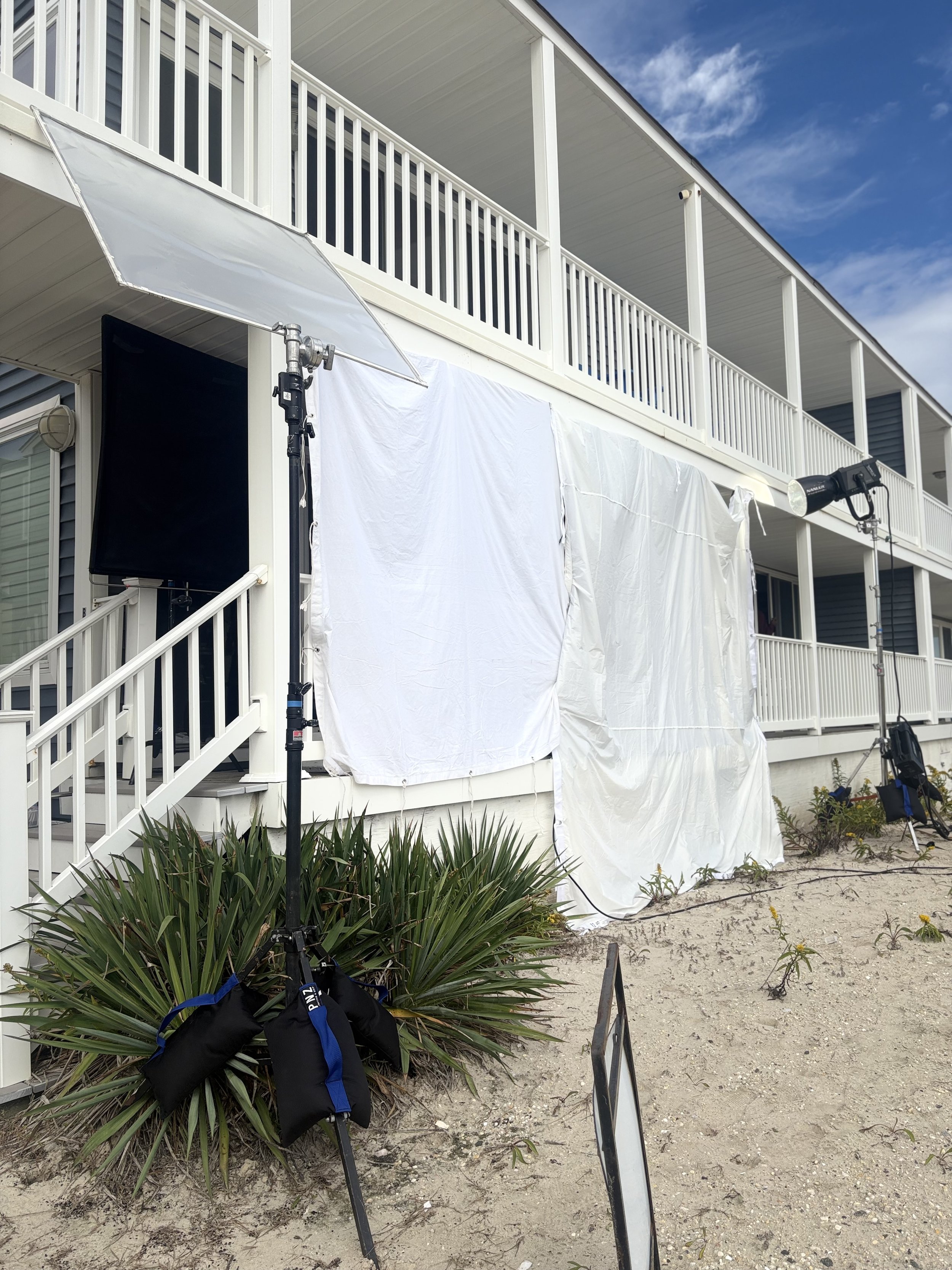 A photo of a two-story beach house with white railing and a large deck, under a partly cloudy sky. Film equipment, including a large softbox light, a spotlight, and a black and blue bag, are set up outside the house on the sandy ground.