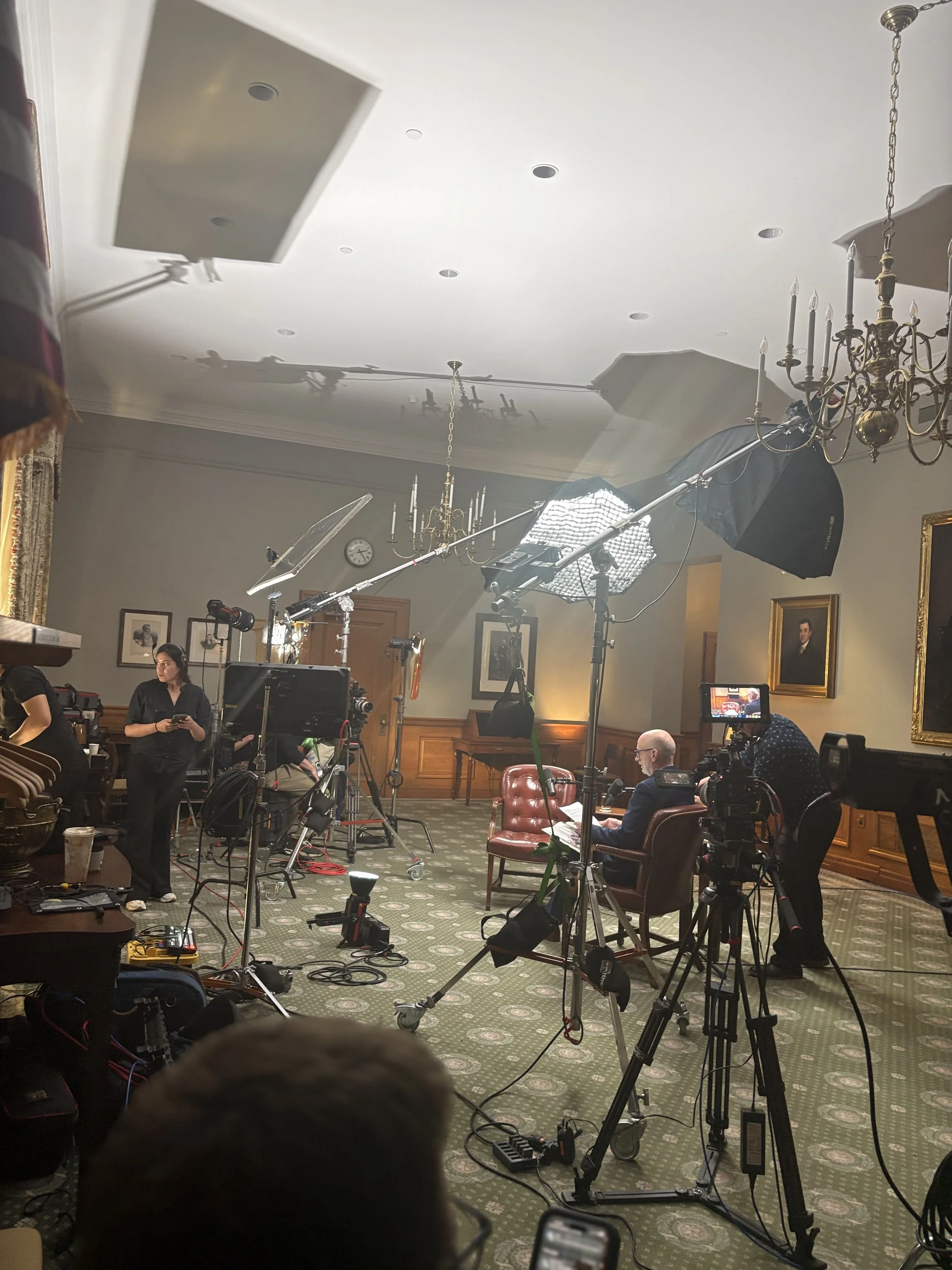 Interior of a room used for filming or photography, with professional lighting and camera equipment set up. There are people working amidst portraits hanging on the wall.