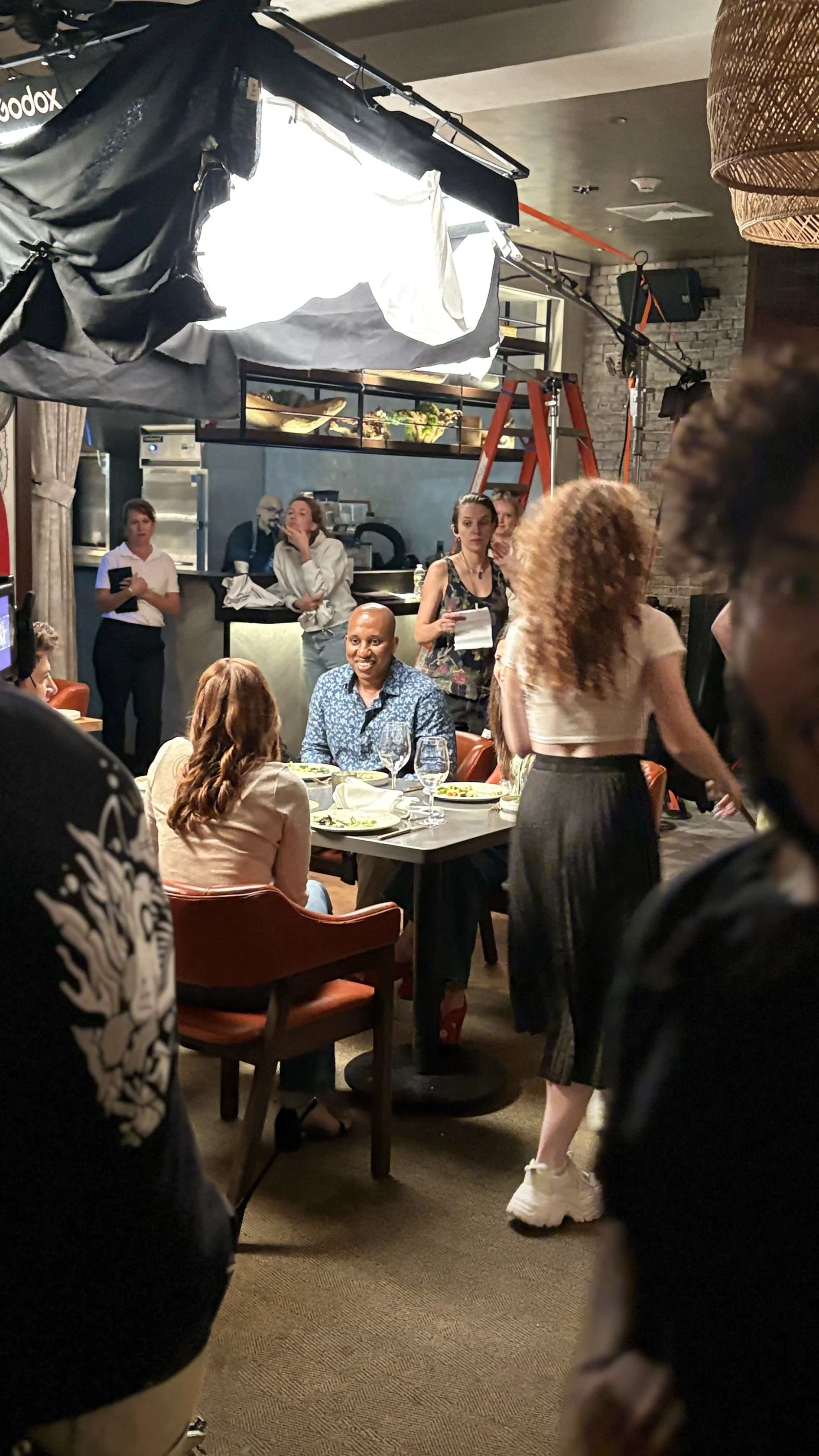 People dining and socializing in a restaurant with a kitchen visible in the background. The scene is lively with some individuals standing and others seated at tables, engaging in conversations. Key Grip - Feature Film "Run"