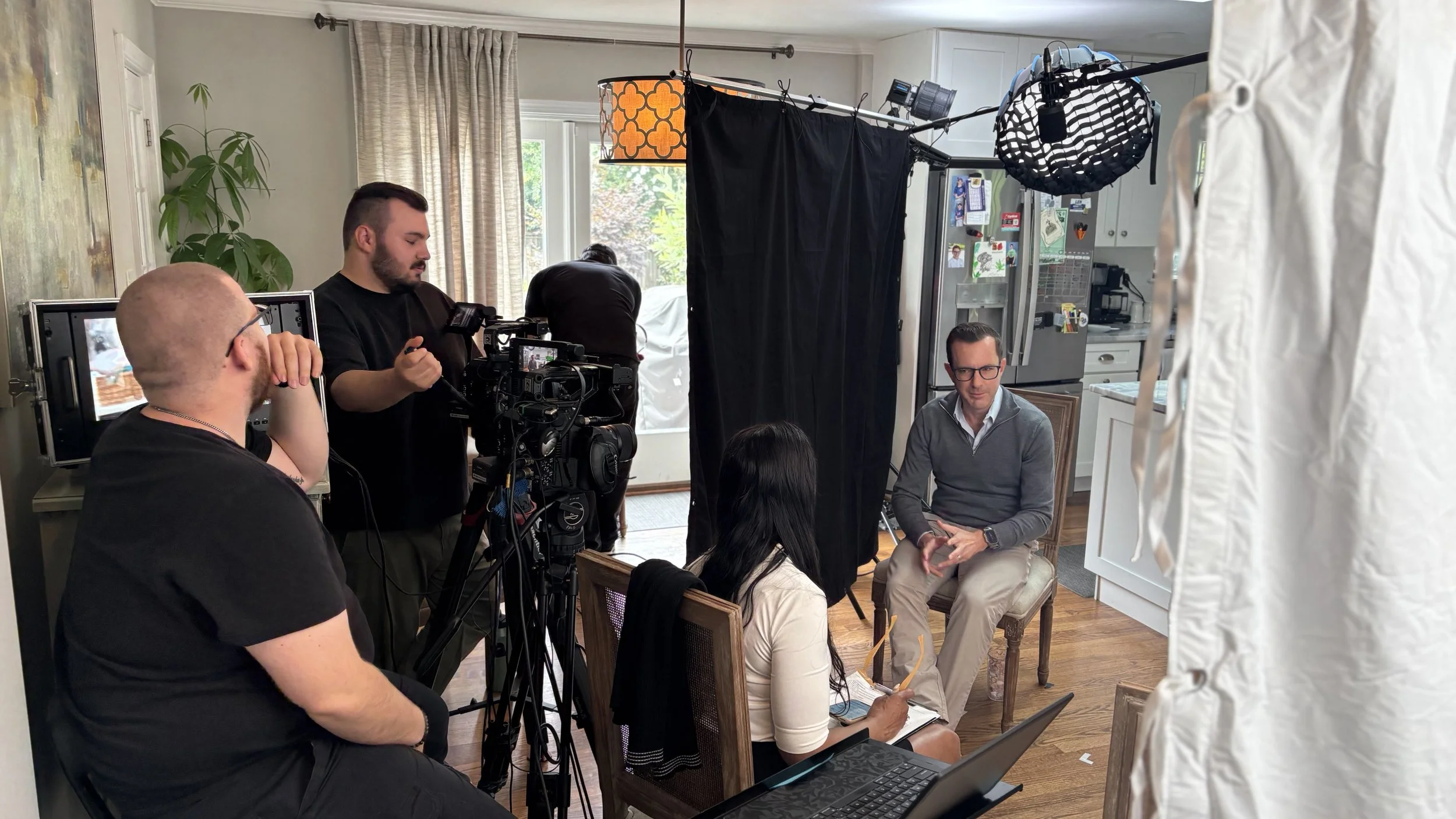 Filming a video interview in a cozy kitchen with two people seated and crew members operating camera equipment and lighting.