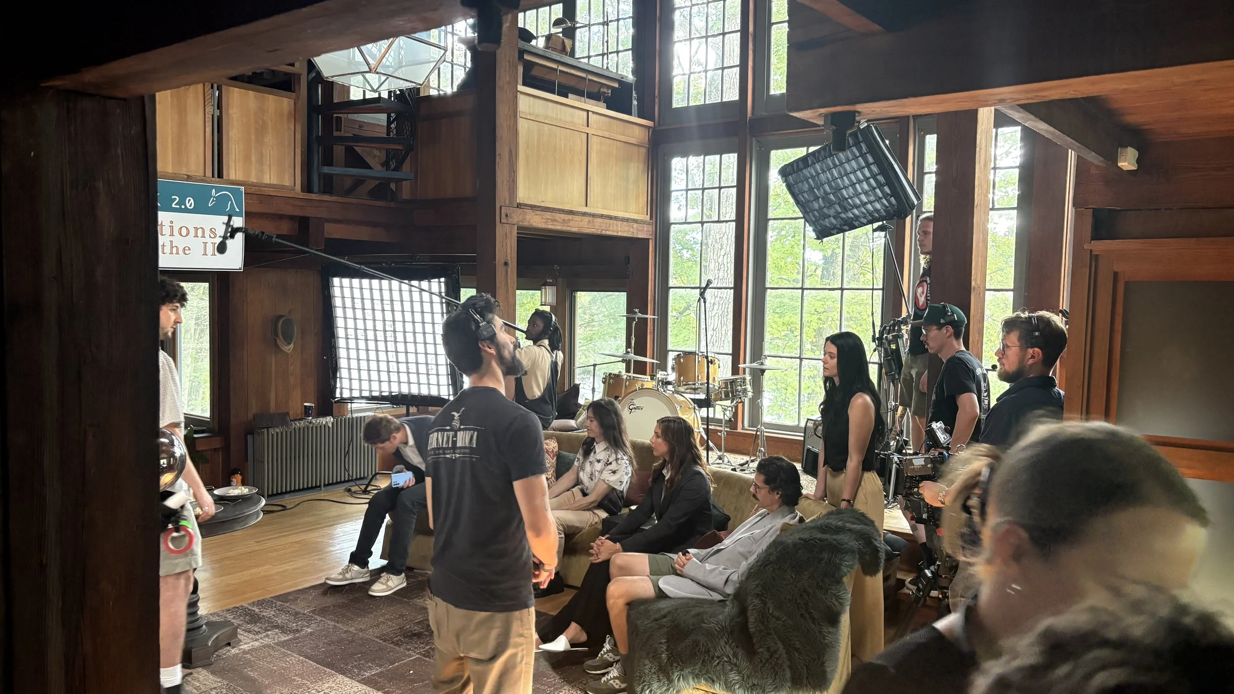 A film crew captures a scene inside a wooden-paneled room with large windows, with multiple crew members and actors, including a woman sitting on a couch and a man with a camera, amid equipment like lights and microphones.
