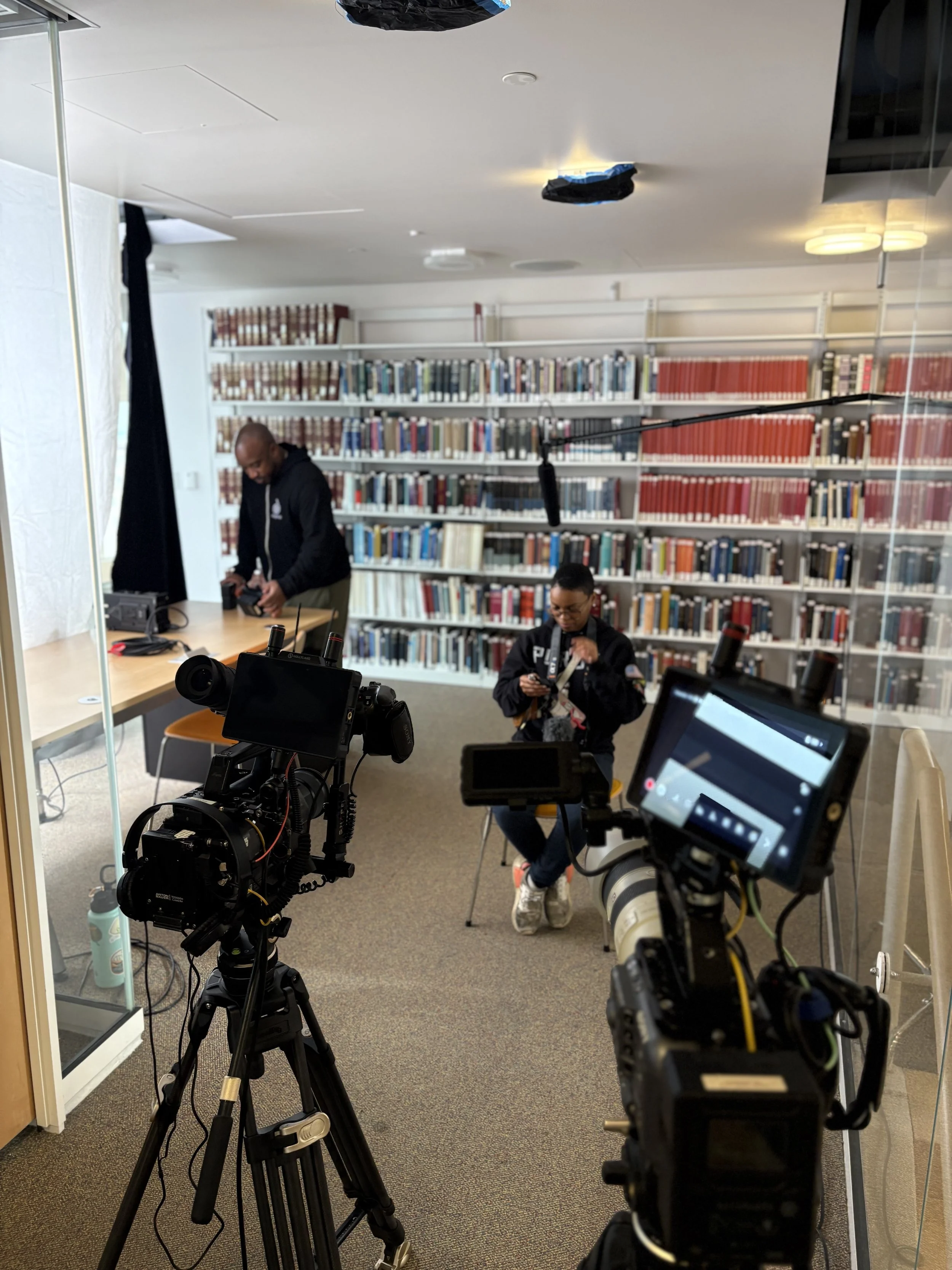 Documentary interview setup: Gaffing and Grip
Two cameras are set up to film or record a scene inside a library . Two people are present: one man standing and working with electronic equipment, and one woman sitting and looking at her phone.