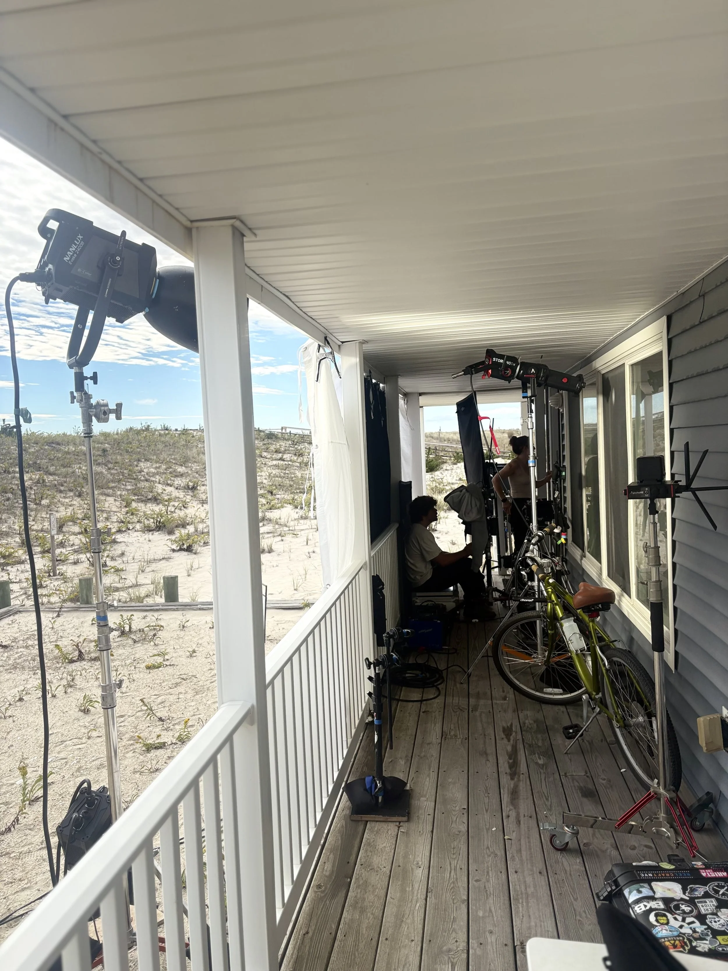 A covered porch area with photography equipment, including lighting, a bicycle, and a storage box, overlooking a sandy beach with some vegetation and a cloudy sky in the background.