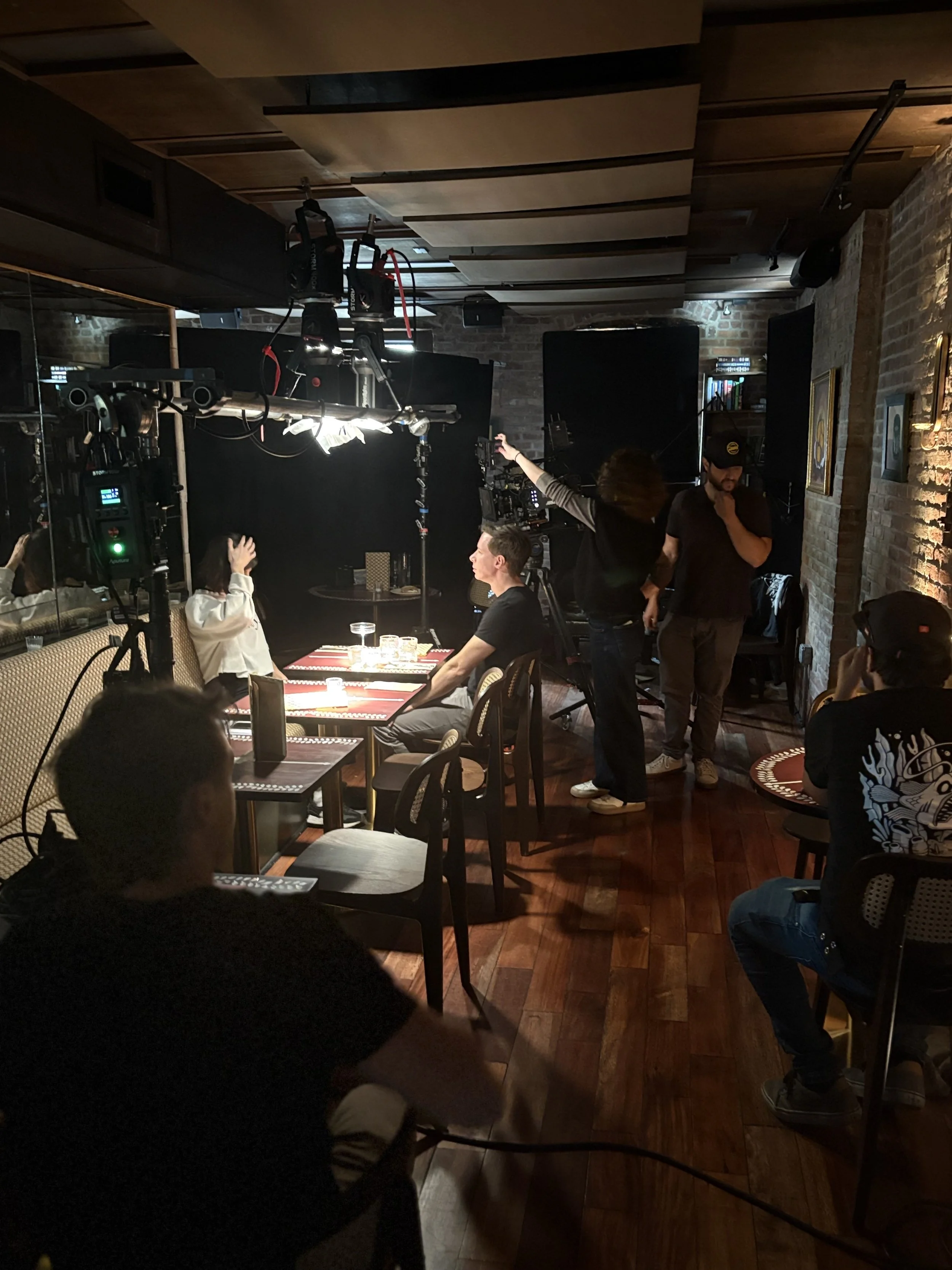 Special Camera Rigging: A behind-the-scenes view of film production in a dimly lit restaurant or cafe setting with crew members, camera equipment, and patrons at tables.