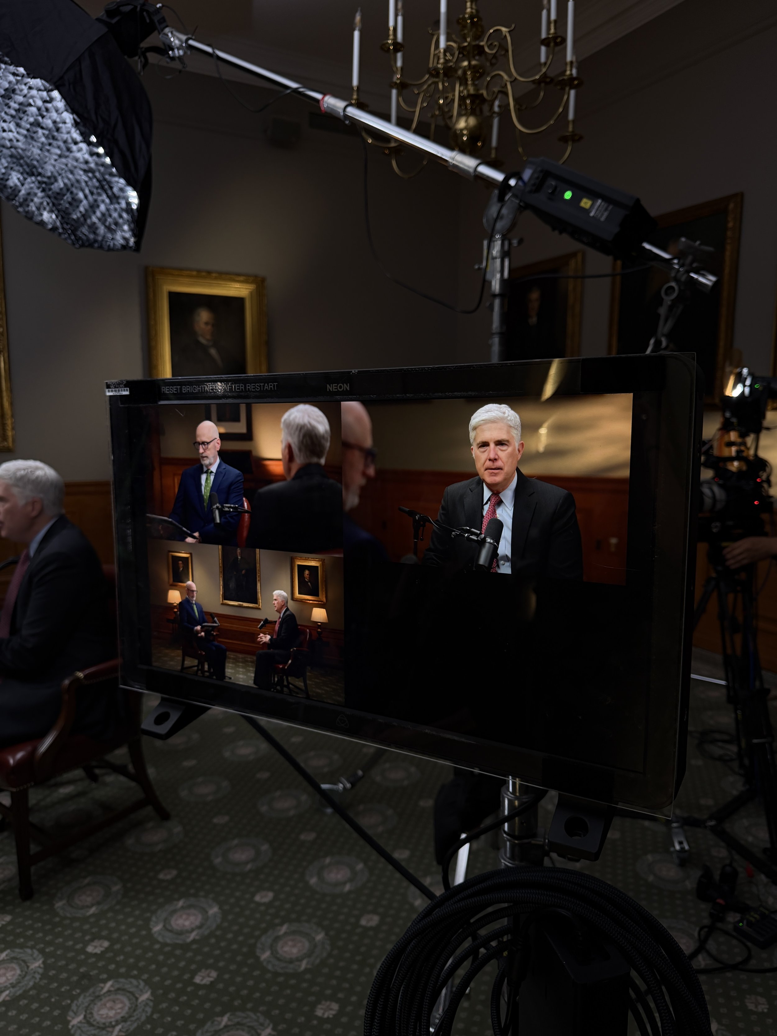 Supreme Court Justice Interview:  A television screen displays an interview with two men seated in a wood-paneled room, surrounded by framed portraits and a brass chandelier.