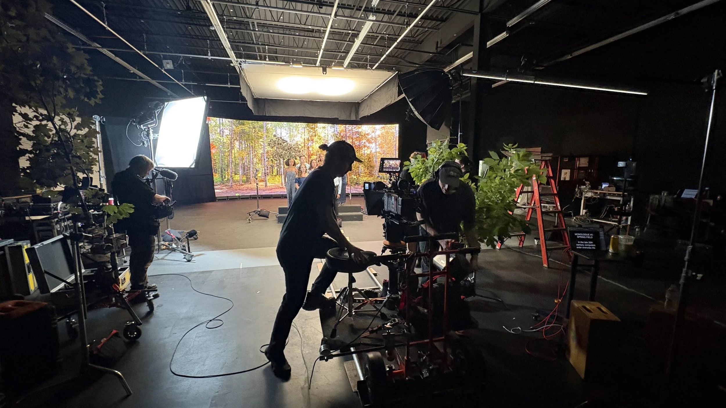 Behind the scenes of a video shoot, with crew members working with equipment and a large screen displaying a scene of people standing outdoors in a forest with autumn foliage.