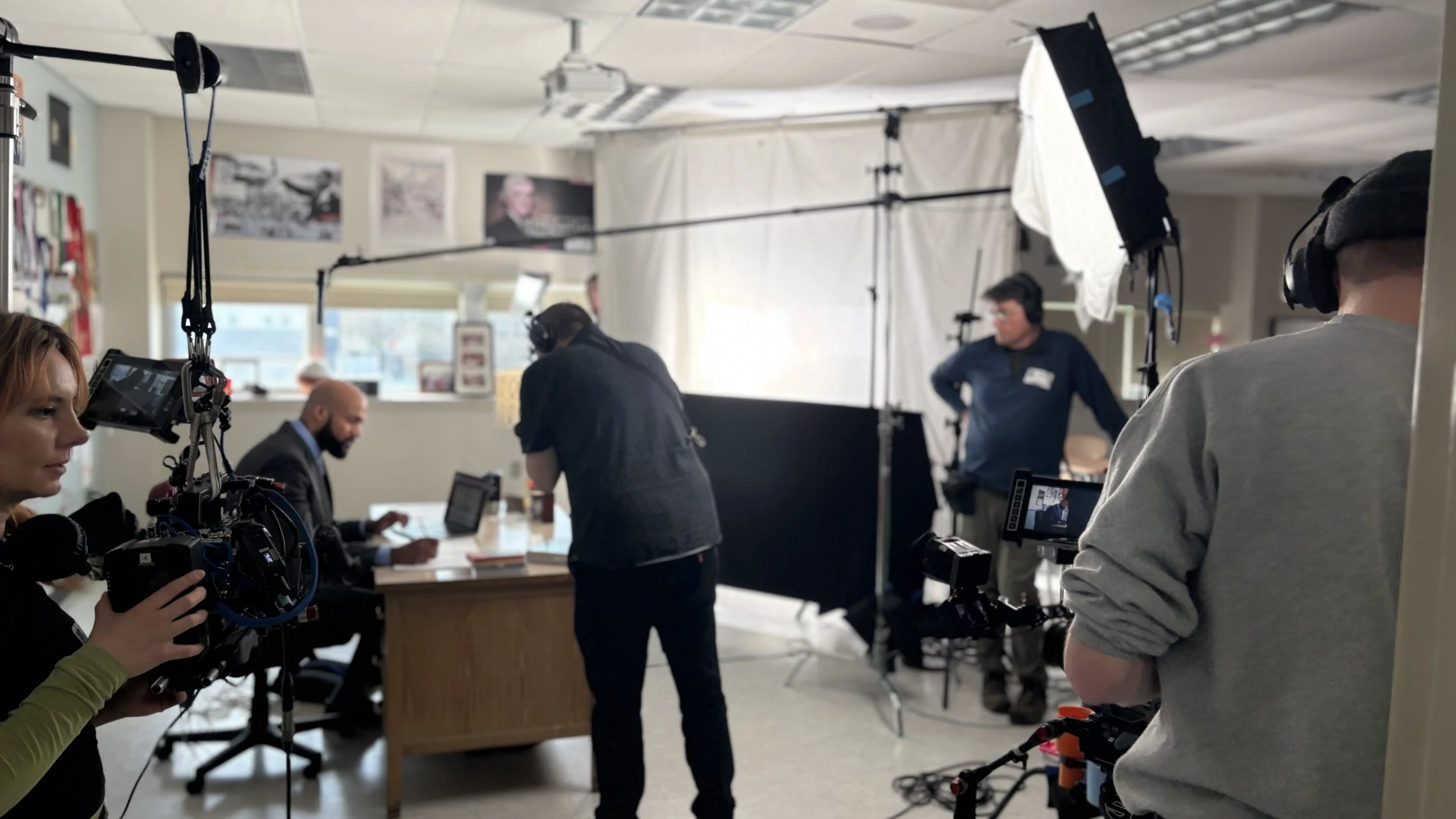 Behind the scenes view of a video or photo shoot, with crew members operating cameras, lighting, and other equipment, and a person seated at a desk.