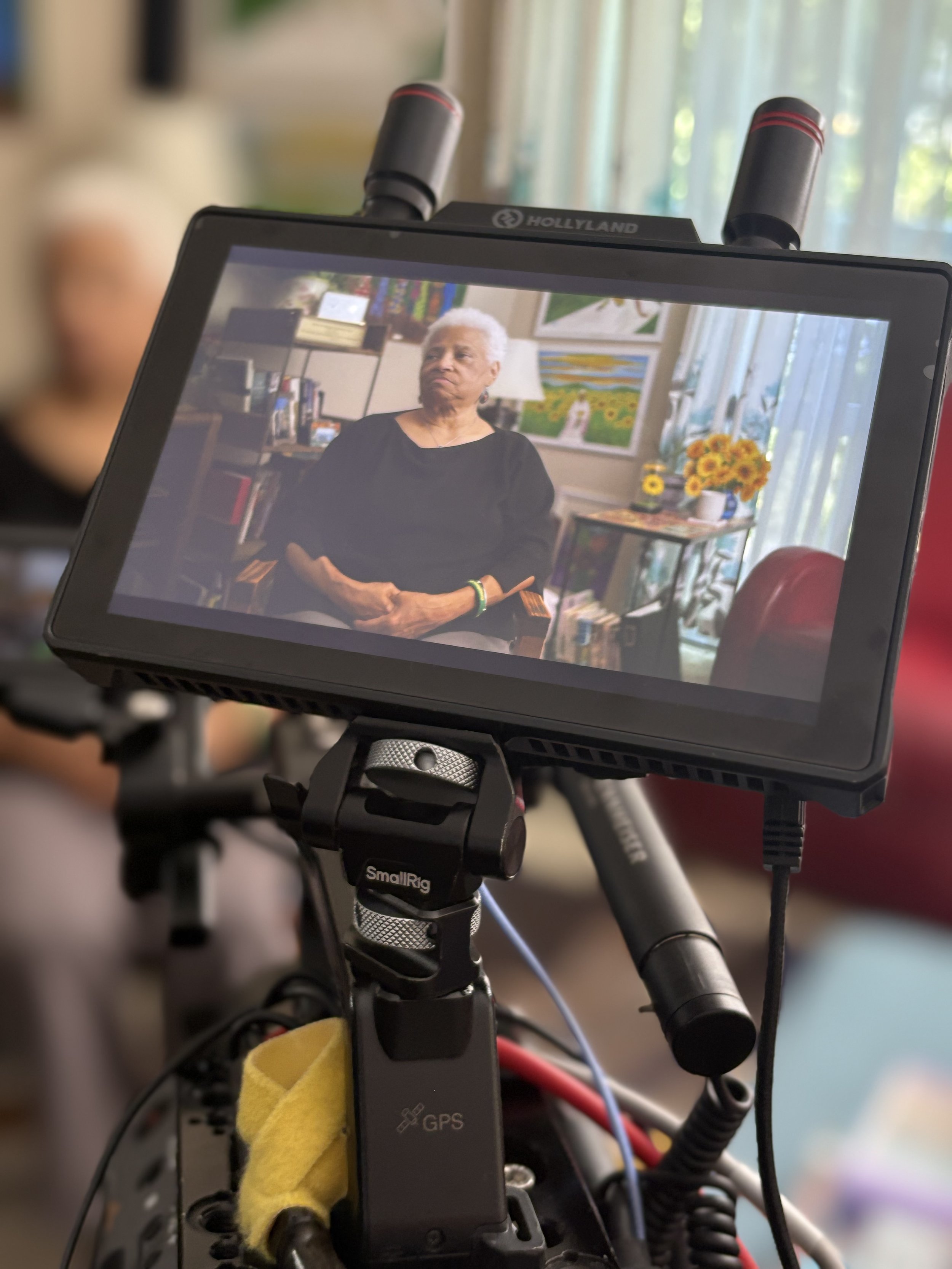 Close-up of a device mounted on a bicycle handlebar displaying a video of an elderly woman with gray hair, wearing a black top, sitting indoors with a colorful background of paintings, flowers, and books.