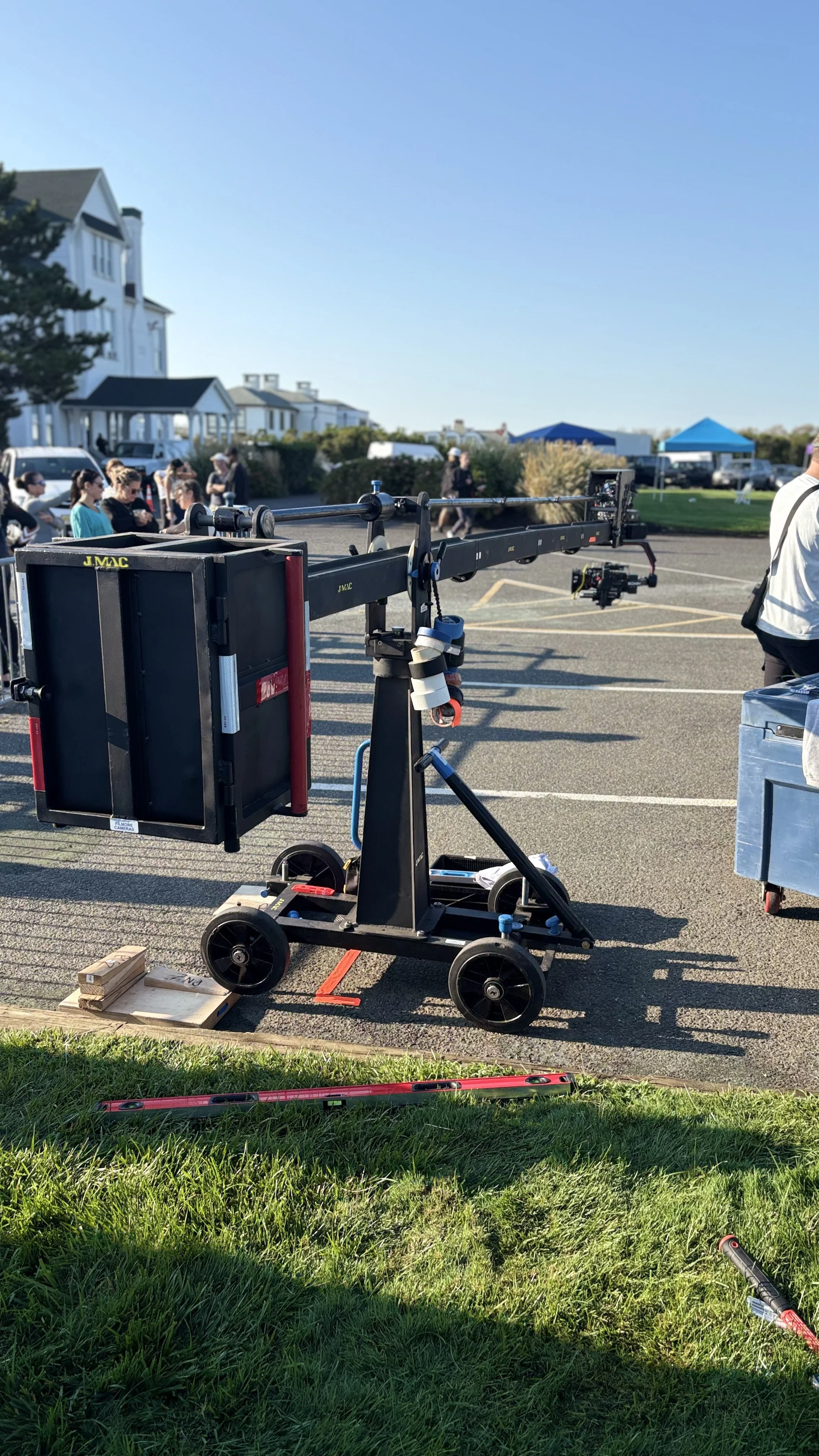Film camera crane setup in an outdoor parking lot with people and tents in the background.