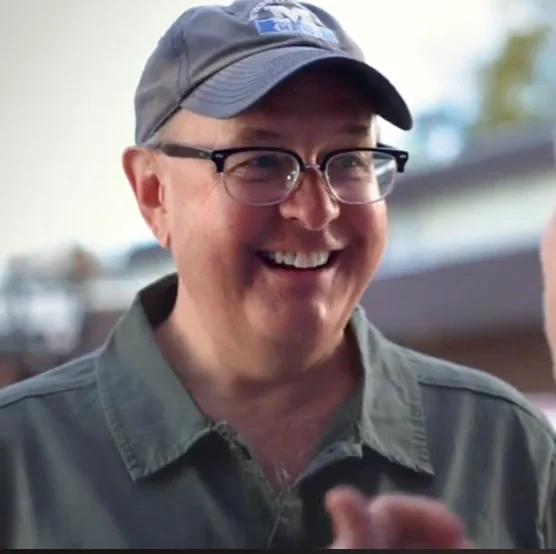 A man with glasses and a cap smiling outdoors.