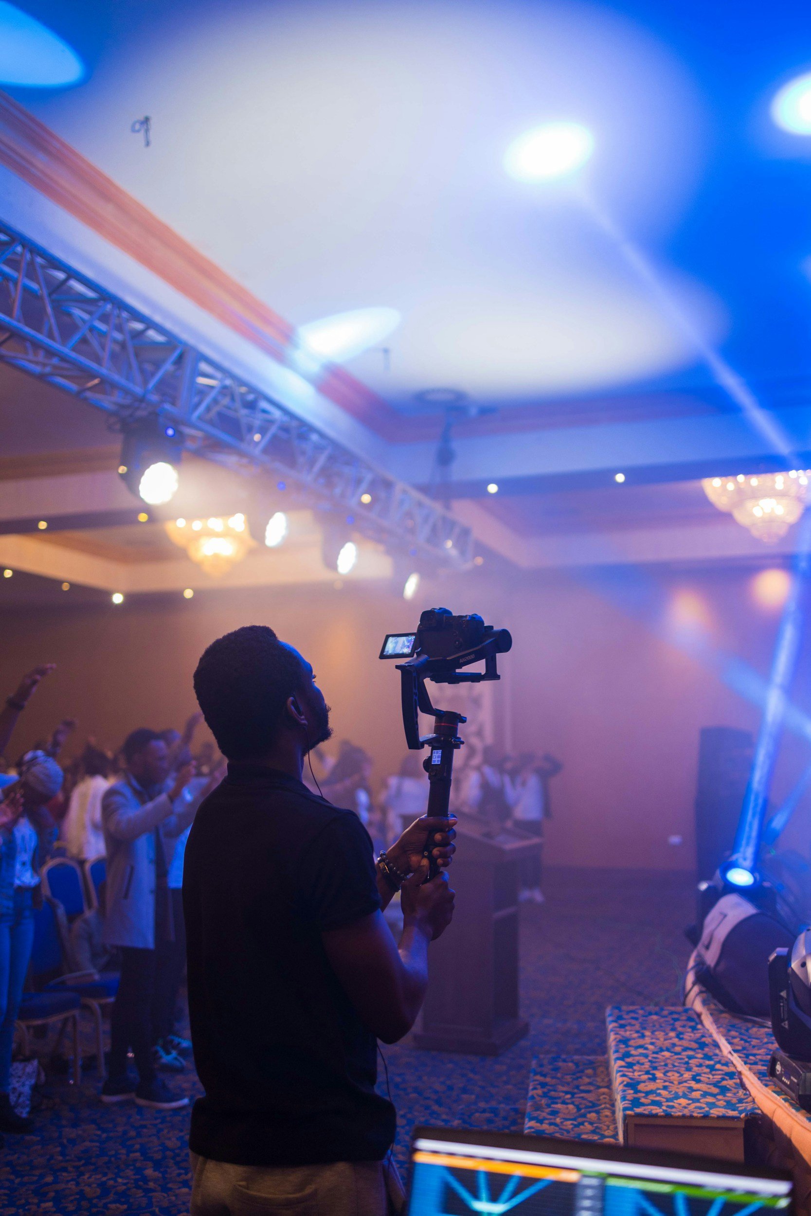 A videographer filming a live event in a ballroom with stage lighting, truss rigging, and a crowd in the background.