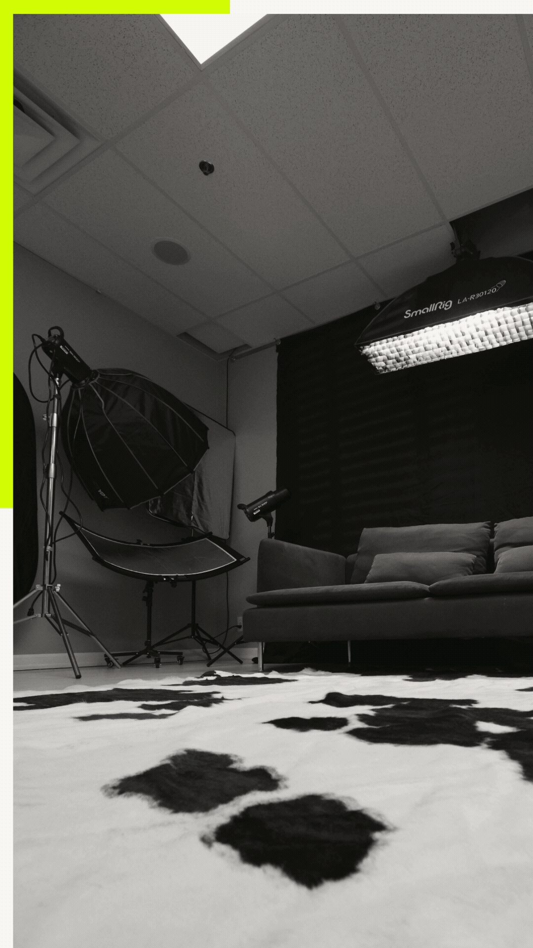 A black and white photo of a photography or video studio with lighting equipment, a sofa, curtains, and a patterned carpet.