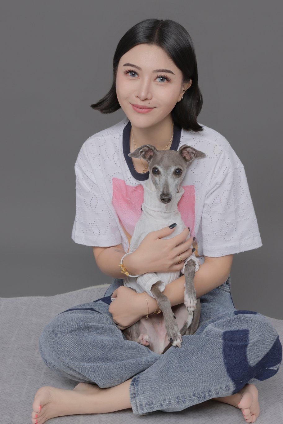 Young woman with short dark hair, sitting cross-legged on a gray surface, holding an Italian Greyhound dog in her lap. She is smiling, wearing a white top with pink design and gray jeans, against a plain gray background.