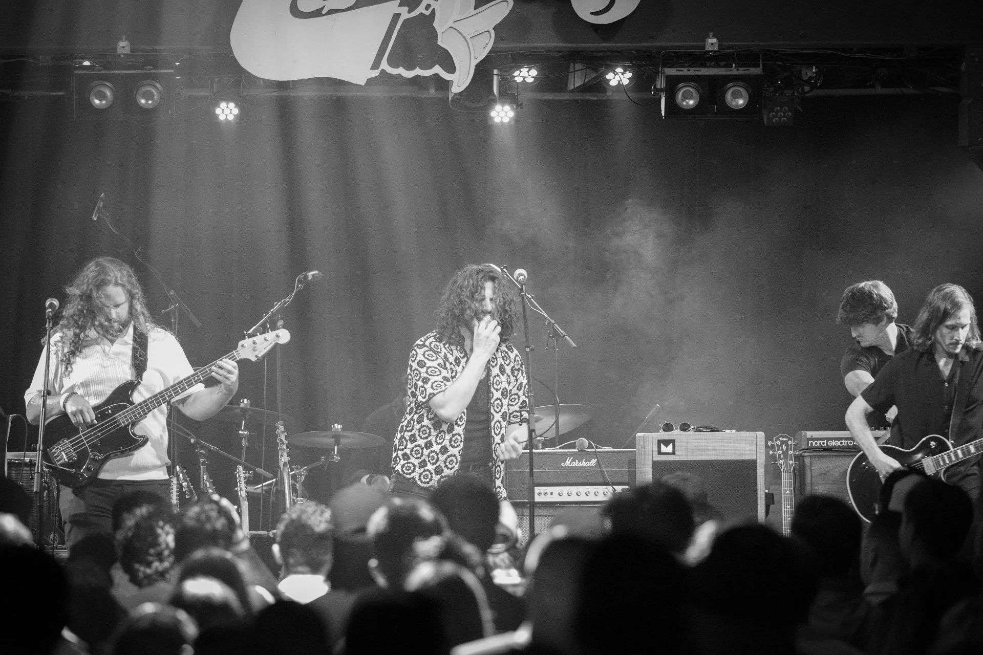 A black-and-white photograph of a live musical performance on stage with a guitarist, a singer, and another musician with a keyboard. The audience is visible in the foreground.