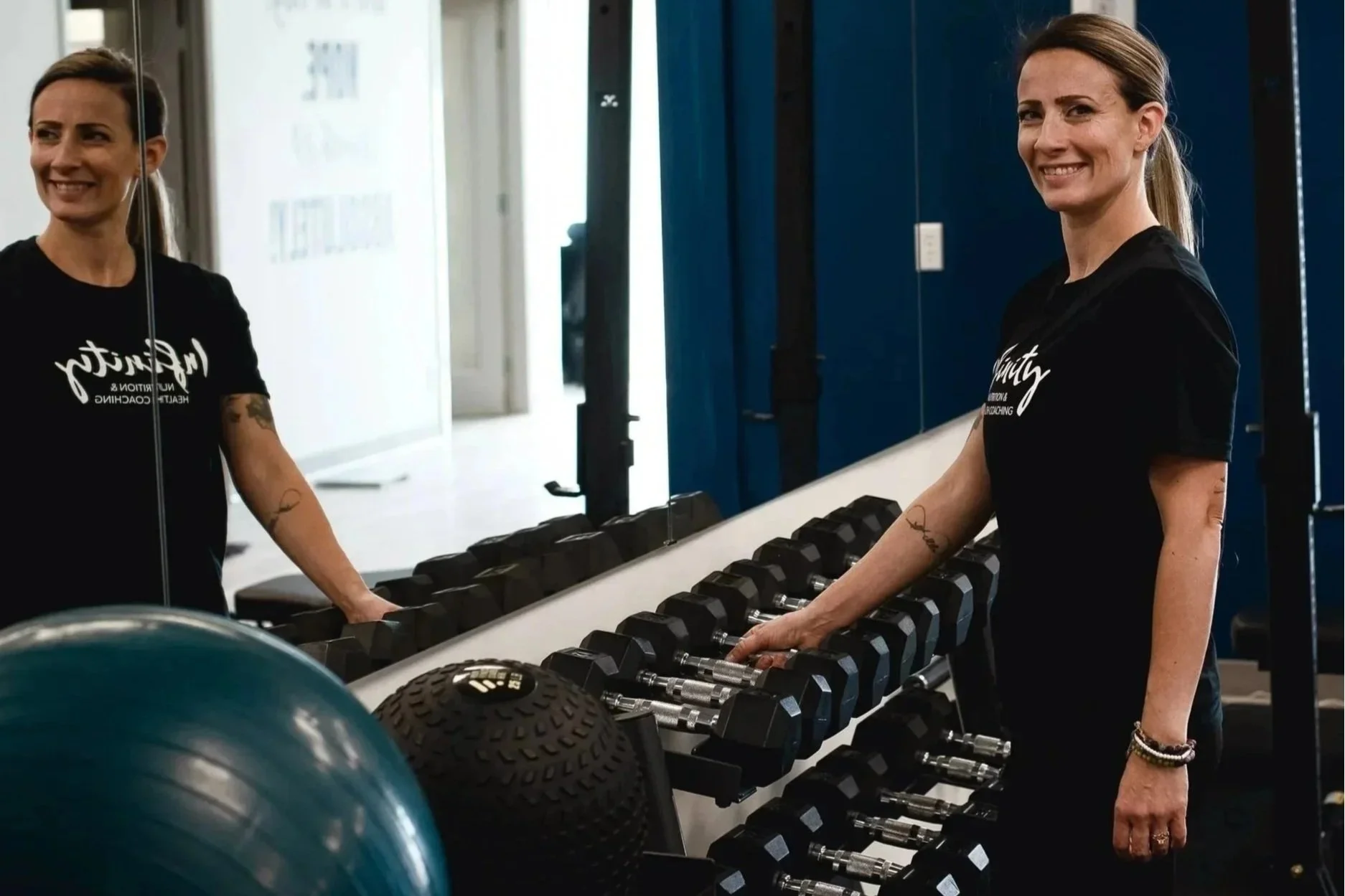 Smiling woman in black T-shirt working with dumbbells at gym, reflected in mirror, with fitness equipment around