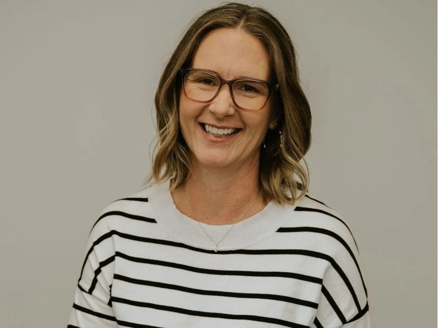 A woman with shoulder-length wavy hair, glasses, and a striped black and white shirt, smiling against a plain gray background.