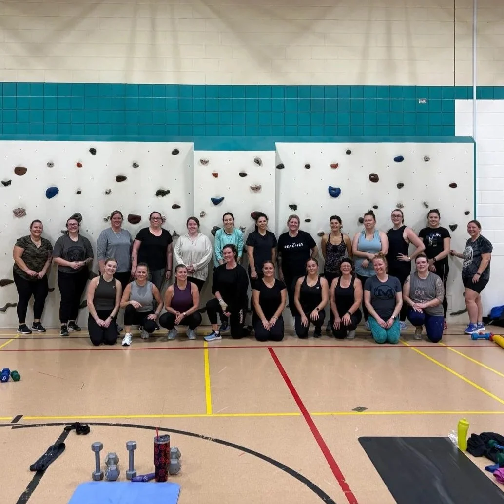 Group of women in workout attire posing in a gymnasium in front of a climbing wall with colorful holds.