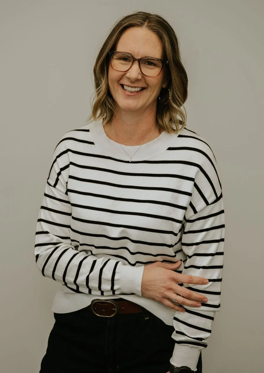 A woman with glasses smiling, wearing a white and black striped sweater, black pants, and a belt, standing against a plain light-colored background.
