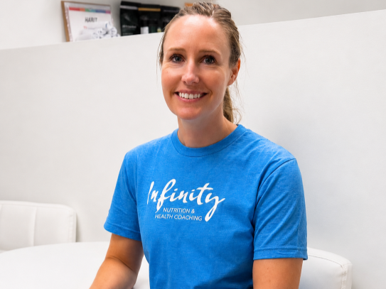 A woman with light brown hair tied back, smiling, wearing a blue t-shirt that reads 'Infnity Nutrition & Health Coaching,' seated indoors against a white wall.