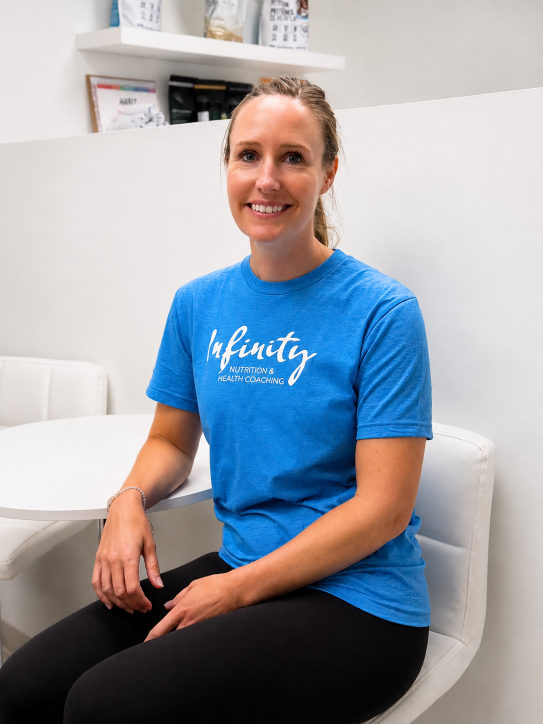 A woman sitting on a white chair in a bright office, wearing a blue t-shirt with "Infinity Nutrition & Health Coaching" written on it, smiling at the camera.