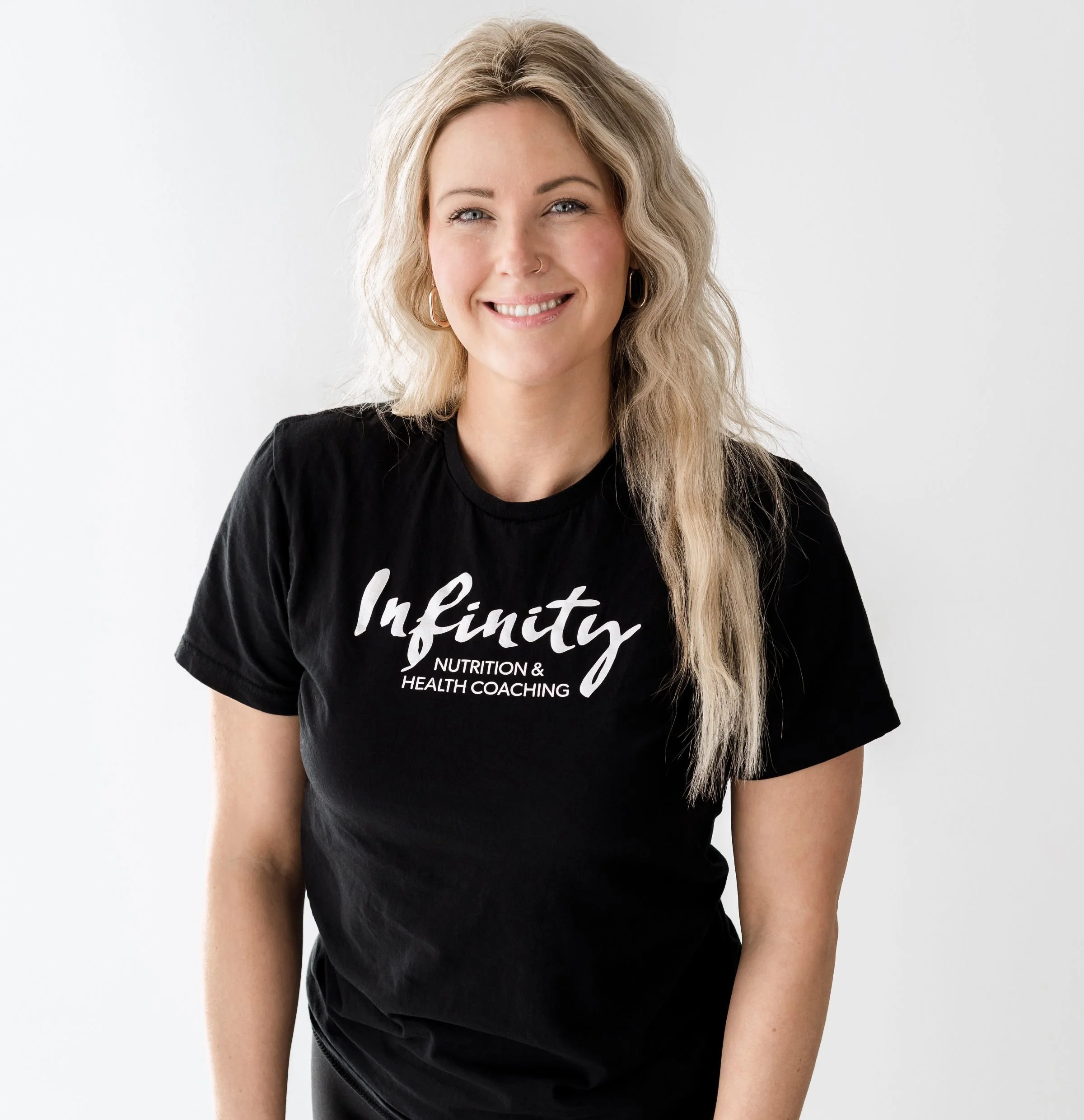 A woman with wavy blonde hair wearing a black t-shirt that says 'Infinity Nutrition & Health Coaching' and smiling, standing against a plain white background.