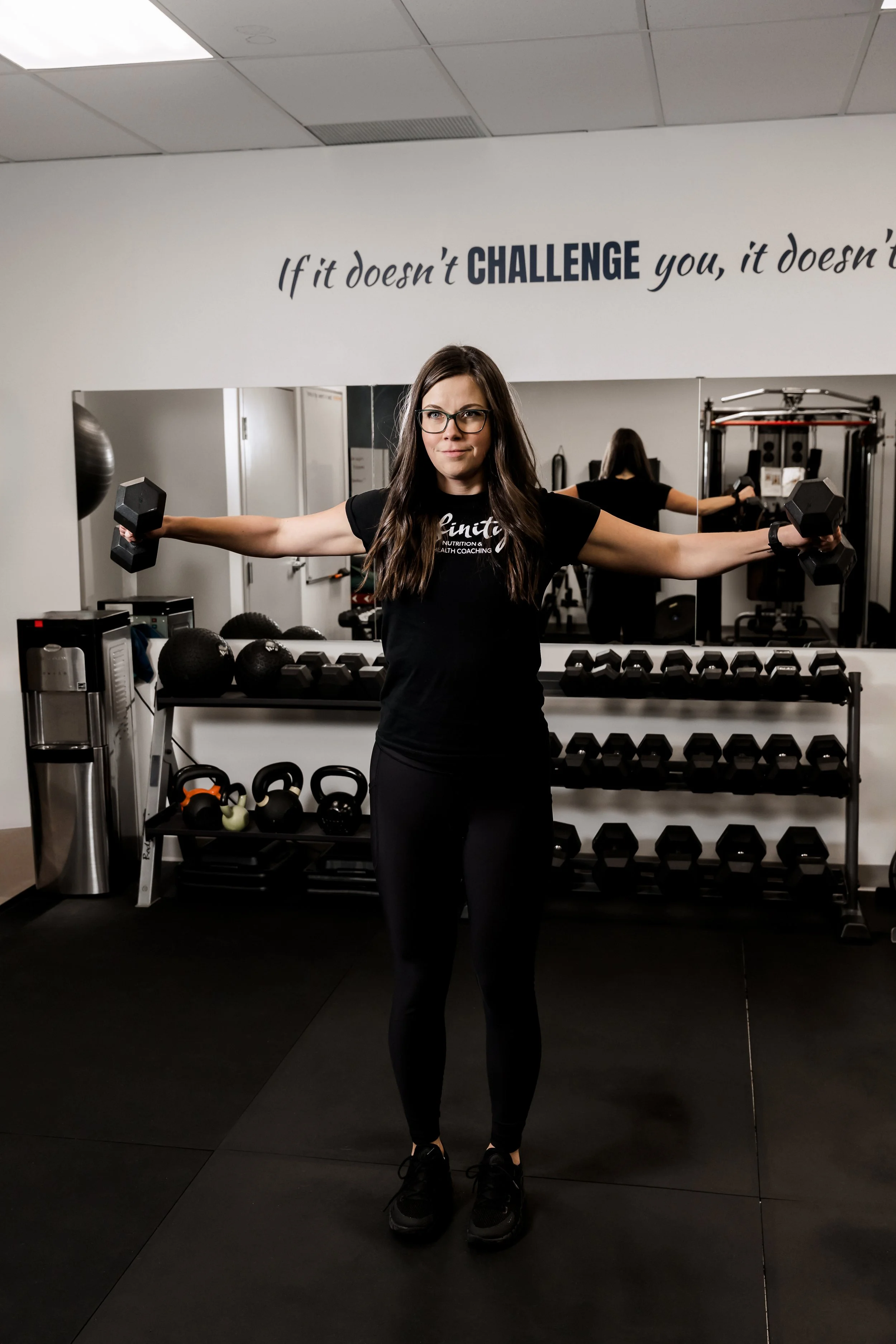 A woman with long dark hair, glasses, and a black workout outfit standing in a gym, holding dumbbells in each hand, with a mirror behind her.