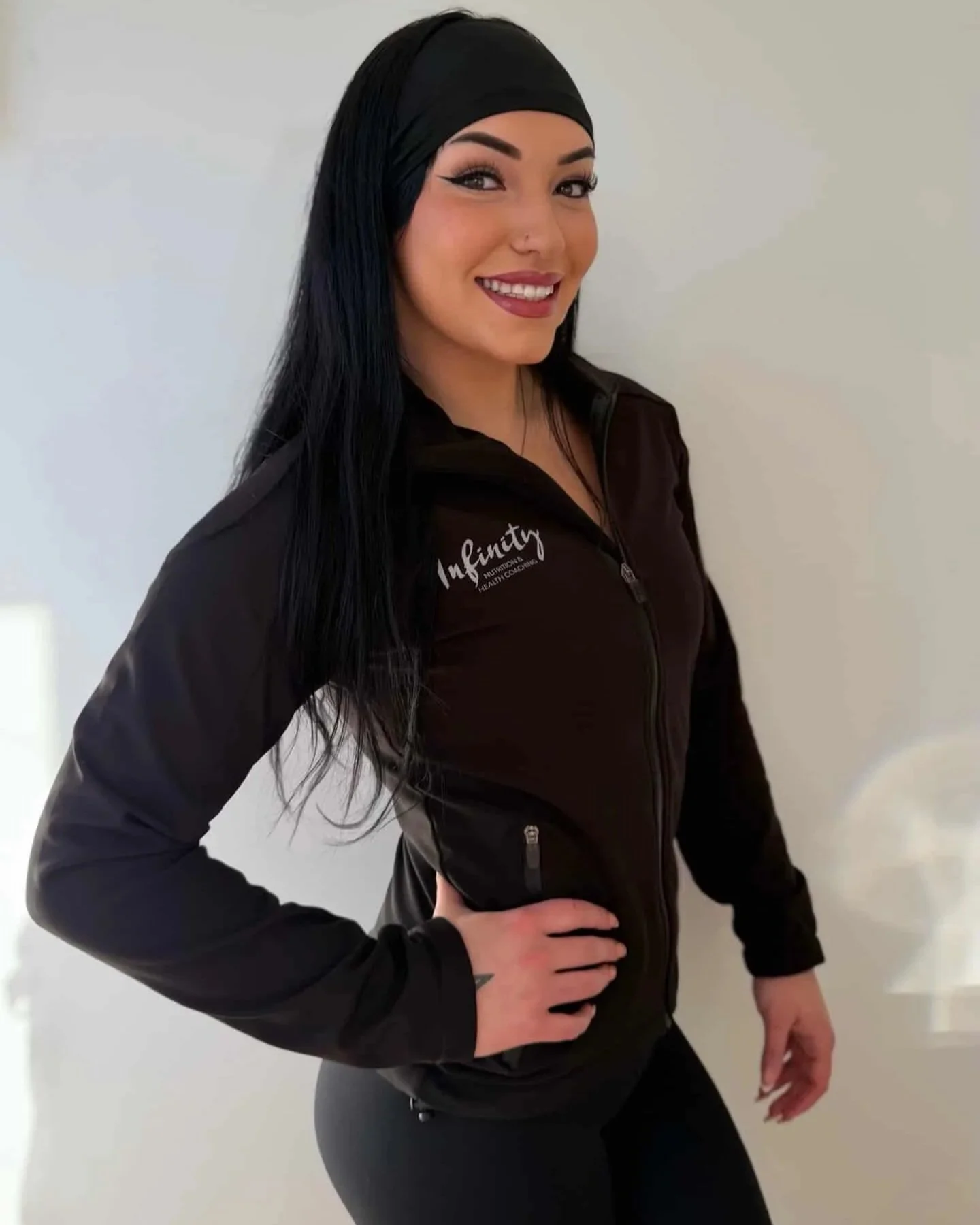 Woman with long black hair wearing a black headband and a black zip-up jacket with 'Infinty' logo, smiling, standing against a white background.