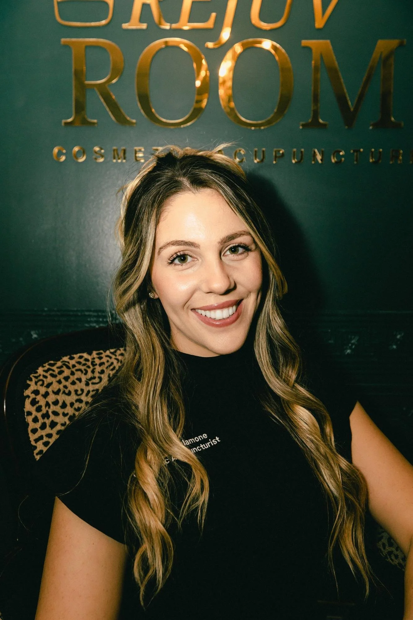 A woman with long wavy blonde hair smiling, sitting in front of a dark green wall with gold lettering that reads 'BEJOV ROOM' and smaller text below. She has light skin, is wearing a black top, and has a leopard print accent on the chair behind her.