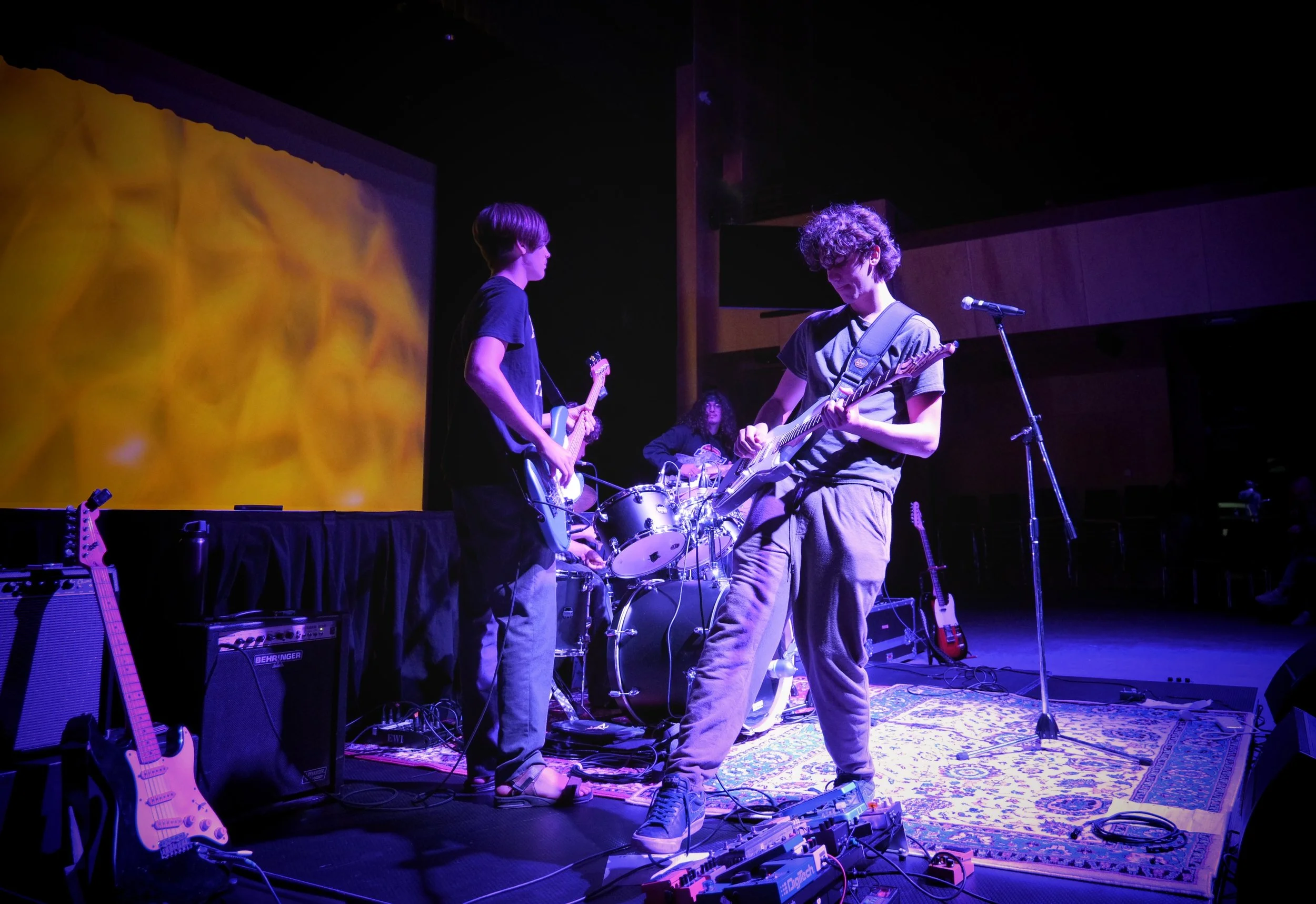 A band of four young musicians performing on stage with guitars, drums, and music equipment, under colorful lighting.