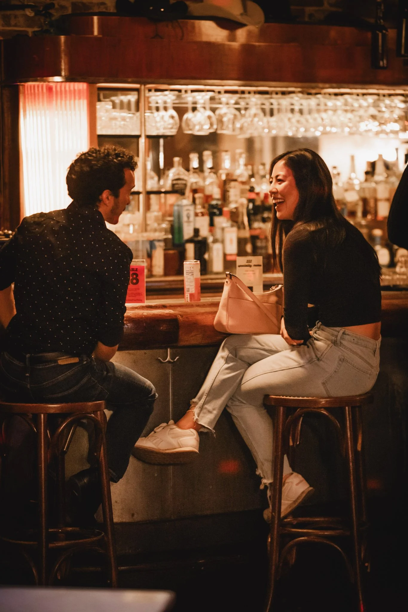 A man and a woman are sitting at a bar, engaged in conversation and laughing. The bar has glasses hanging upside down and bottles of alcohol in the background. The woman is wearing light-colored jeans and a black top, while the man is wearing a dark shirt with white polka dots.