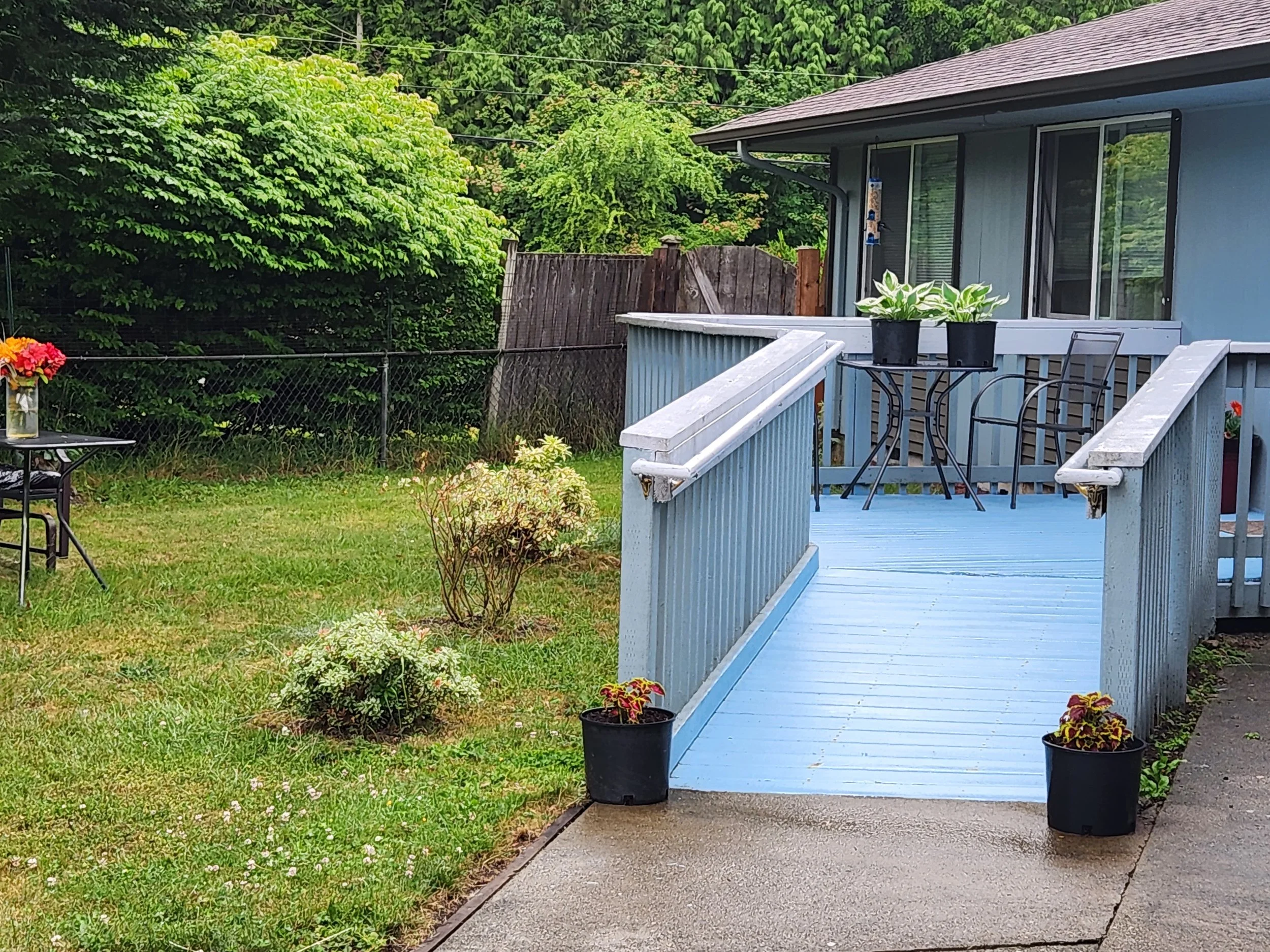 A blue wooden porch with potted plants and outdoor chairs, overlooking a grassy yard enclosed by a fence, with trees in the background.