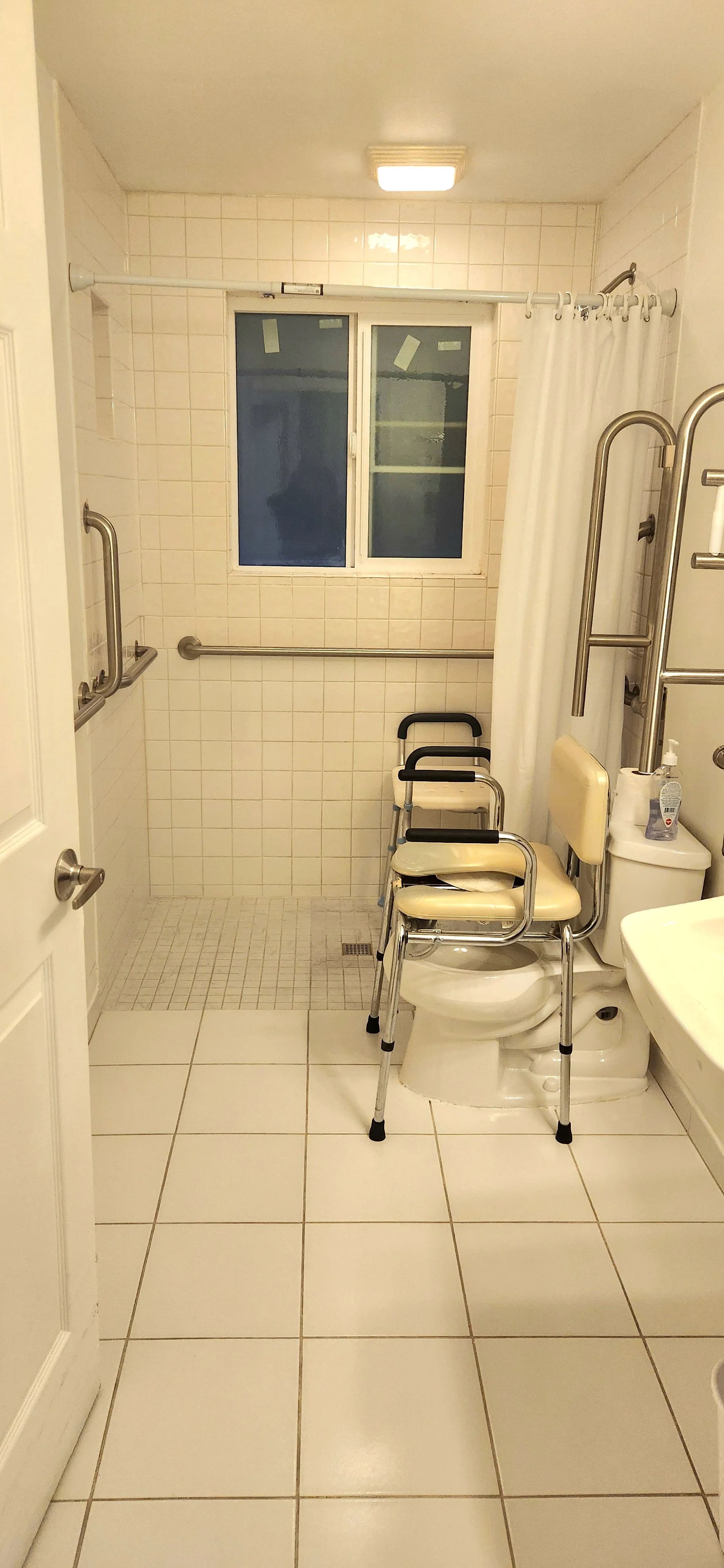 A bathroom with white tiled walls and floor, a small window above, a shower area with grab bars, a white curtain, a toilet with a beige seat, a chair placed over the toilet, hand sanitizer, and a sink.