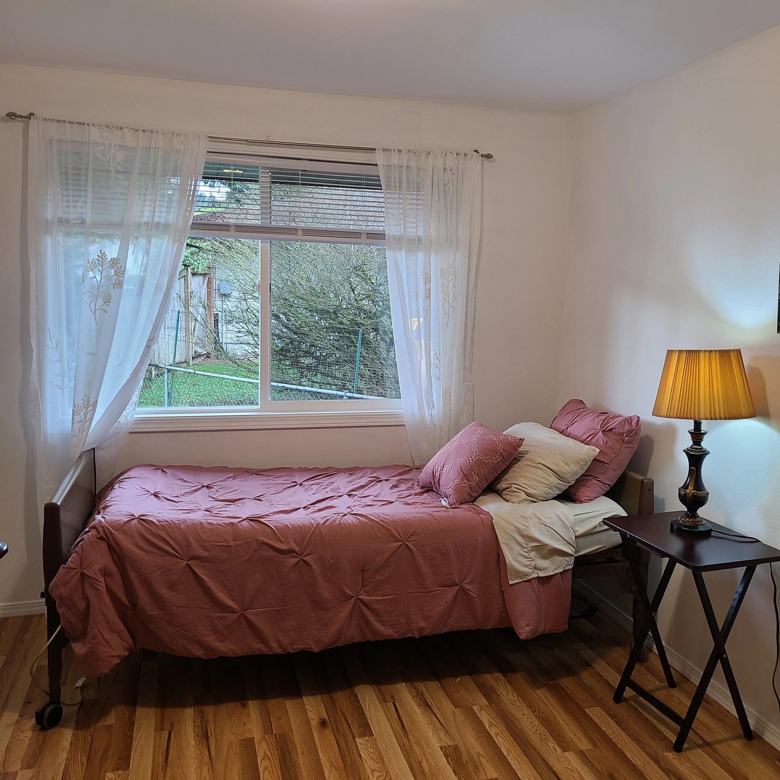A bedroom with a single bed covered in a pink bedspread and pillows, a wooden side table with a lamp, a window with white curtains, and wooden flooring.