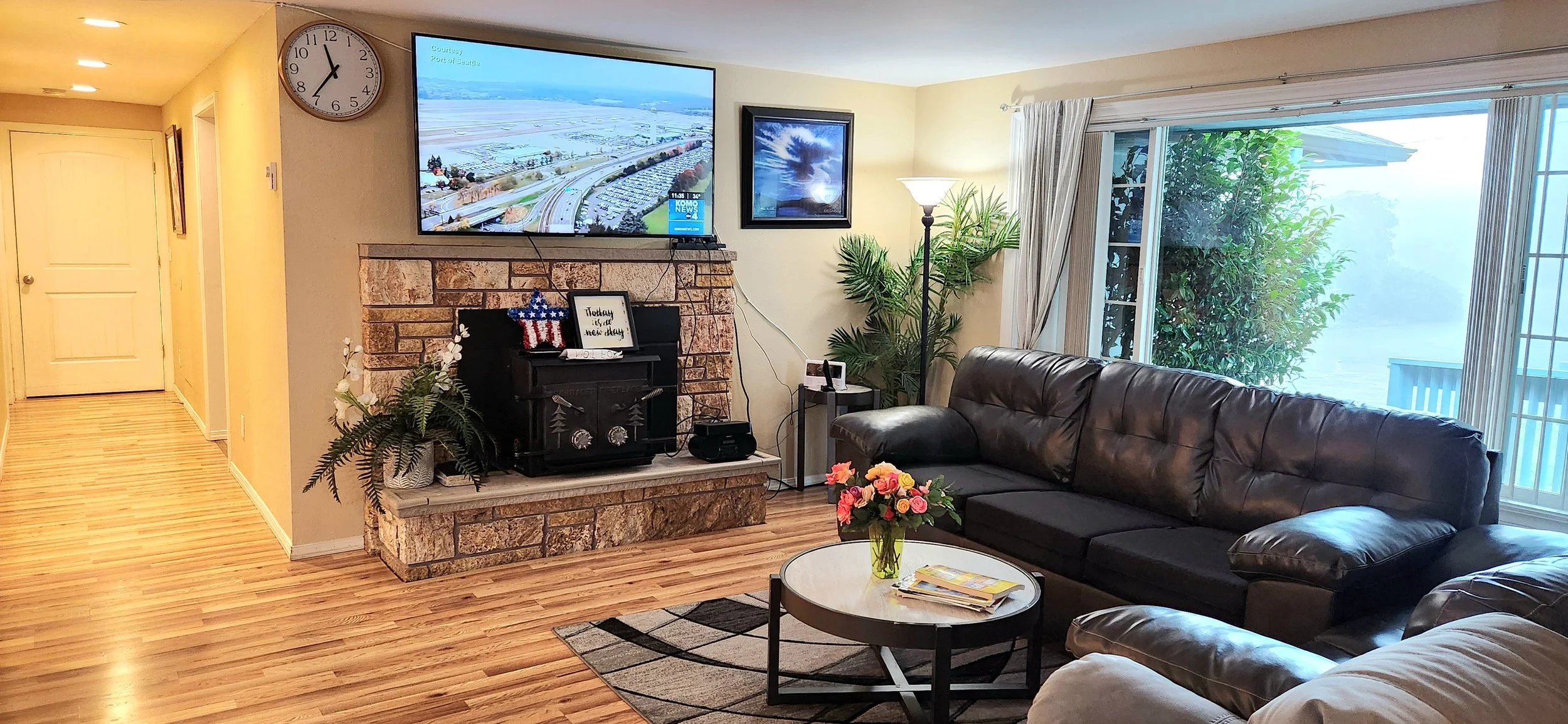 Living room with a brick fireplace, a mounted flat-screen TV, a black leather sofa, a glass coffee table with a bouquet of pink and peach roses, indoor plants, and large sliding glass doors with curtains leading outside.