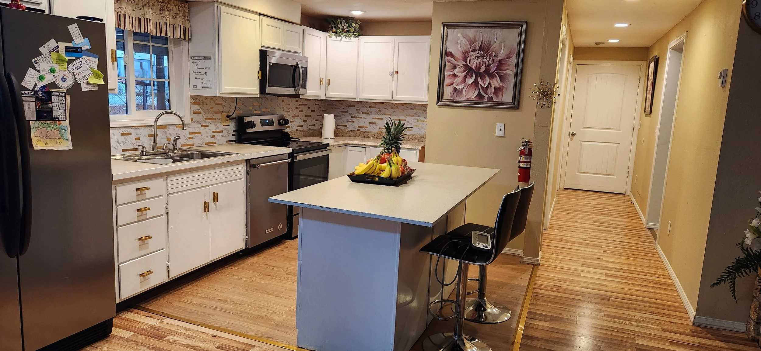A kitchen with white cabinets, a stainless steel refrigerator, a black stove, microwave, and dishwasher. There is a window above the sink, a countertop with a paper towel roll and a pineapple, and a rectangular island with a bowl of bananas and other