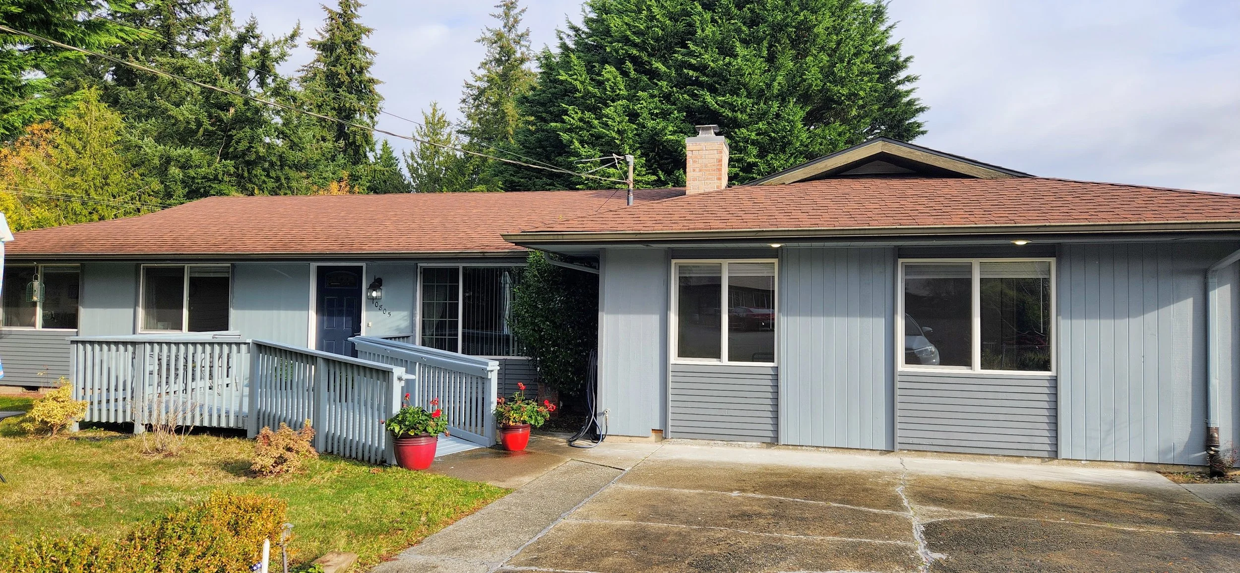 A single-story house with blue siding and a brown shingle roof. There is a small front shrub, two red flower pots with blooms by the entrance, a ramp leading to a side door, and large windows. In the background, tall trees and a cloudy sky are visibl