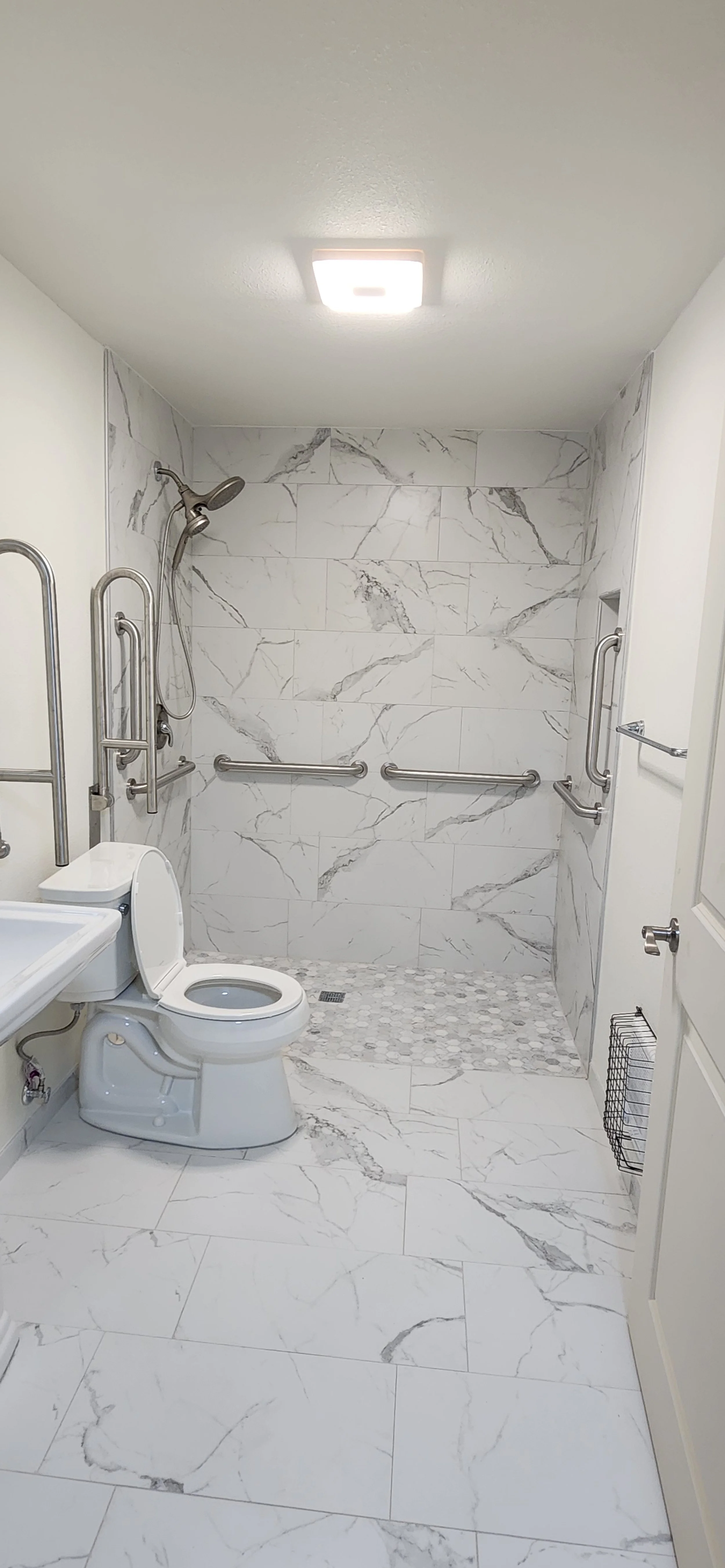 Accessible bathroom with marble tiles, a toilet, grab bars, and a walk-in shower with a small pebble tiled floor.