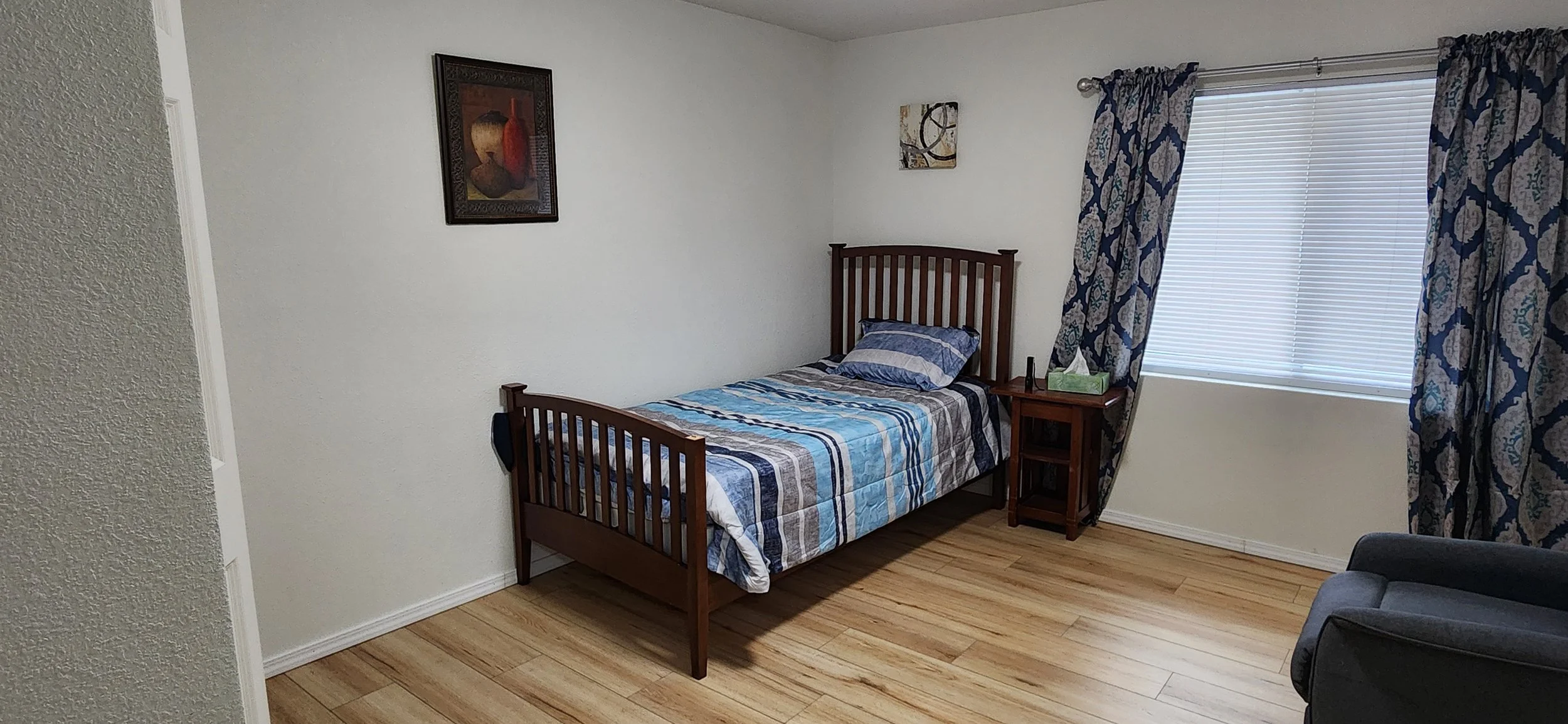 A bedroom with a twin bed, blue and plaid bedspread, wooden headboard and footboard, a side table with a tissue box and a bottle, a window with closed blinds and patterned curtains, a painting on the wall, a small artwork, and part of a gray armchair
