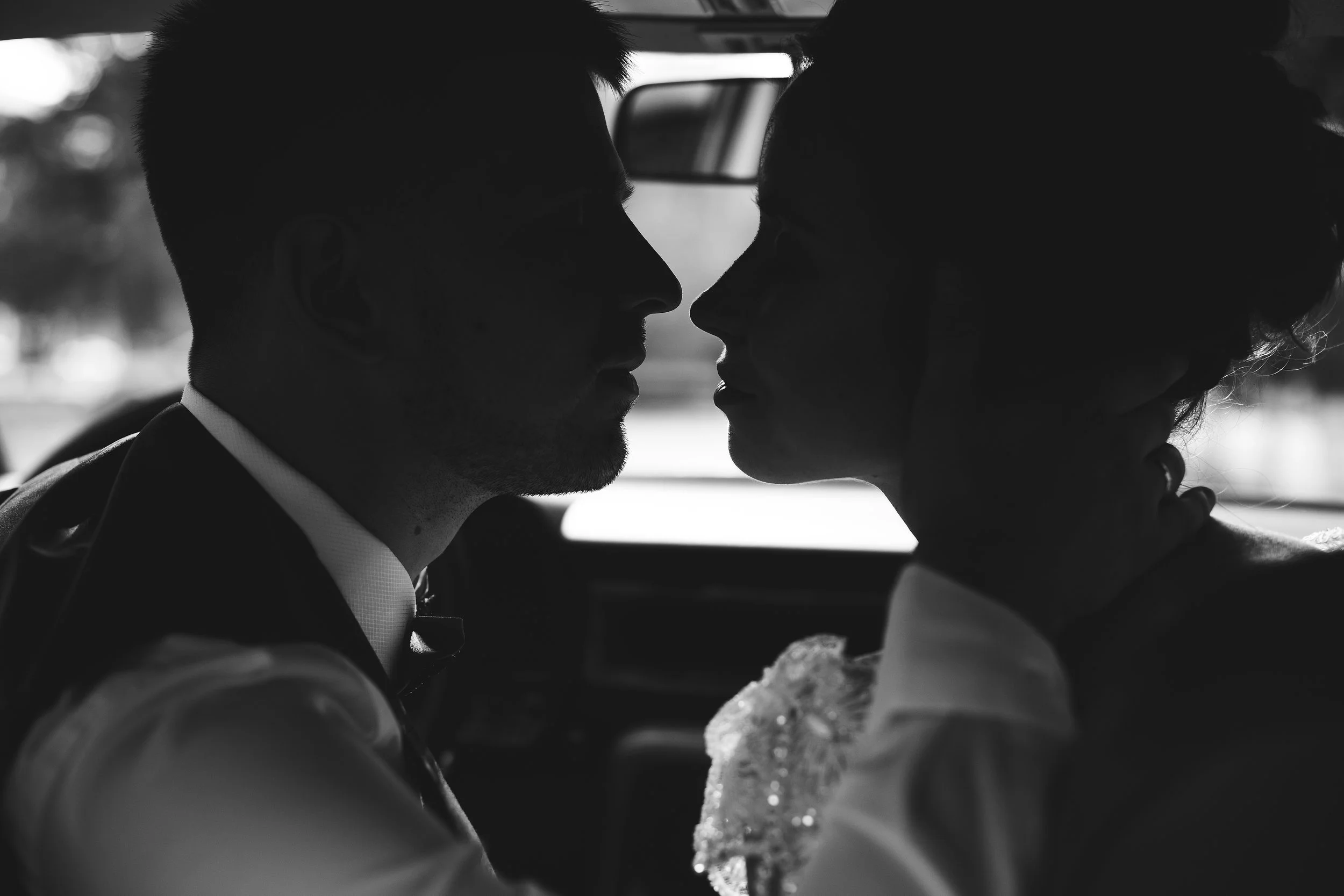 Silhouette of a couple close together, about to kiss inside a car, with a blurred outdoor background.