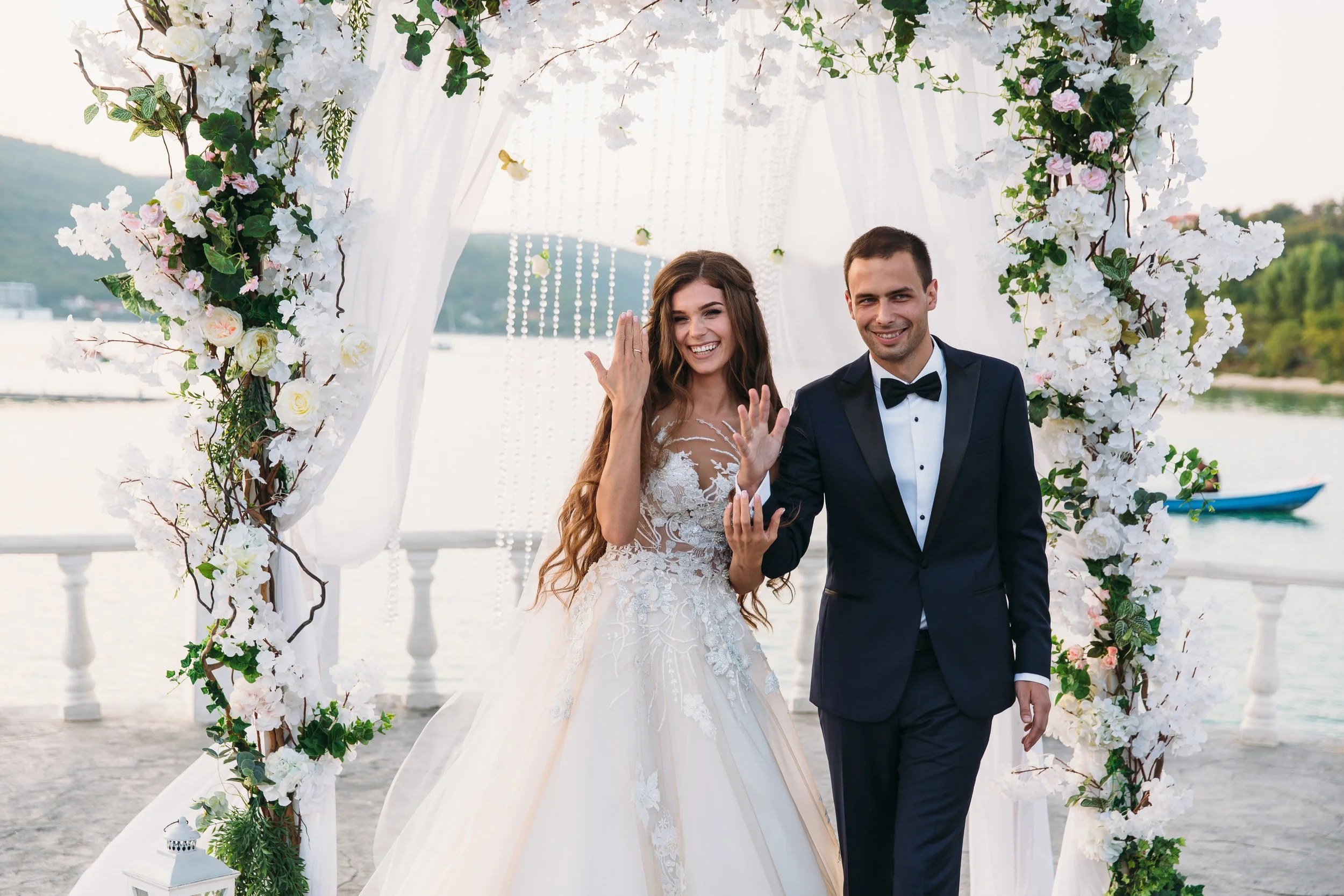 A bride and groom smiling and waving as they walk under a floral arch at an outdoor wedding by a body of water.