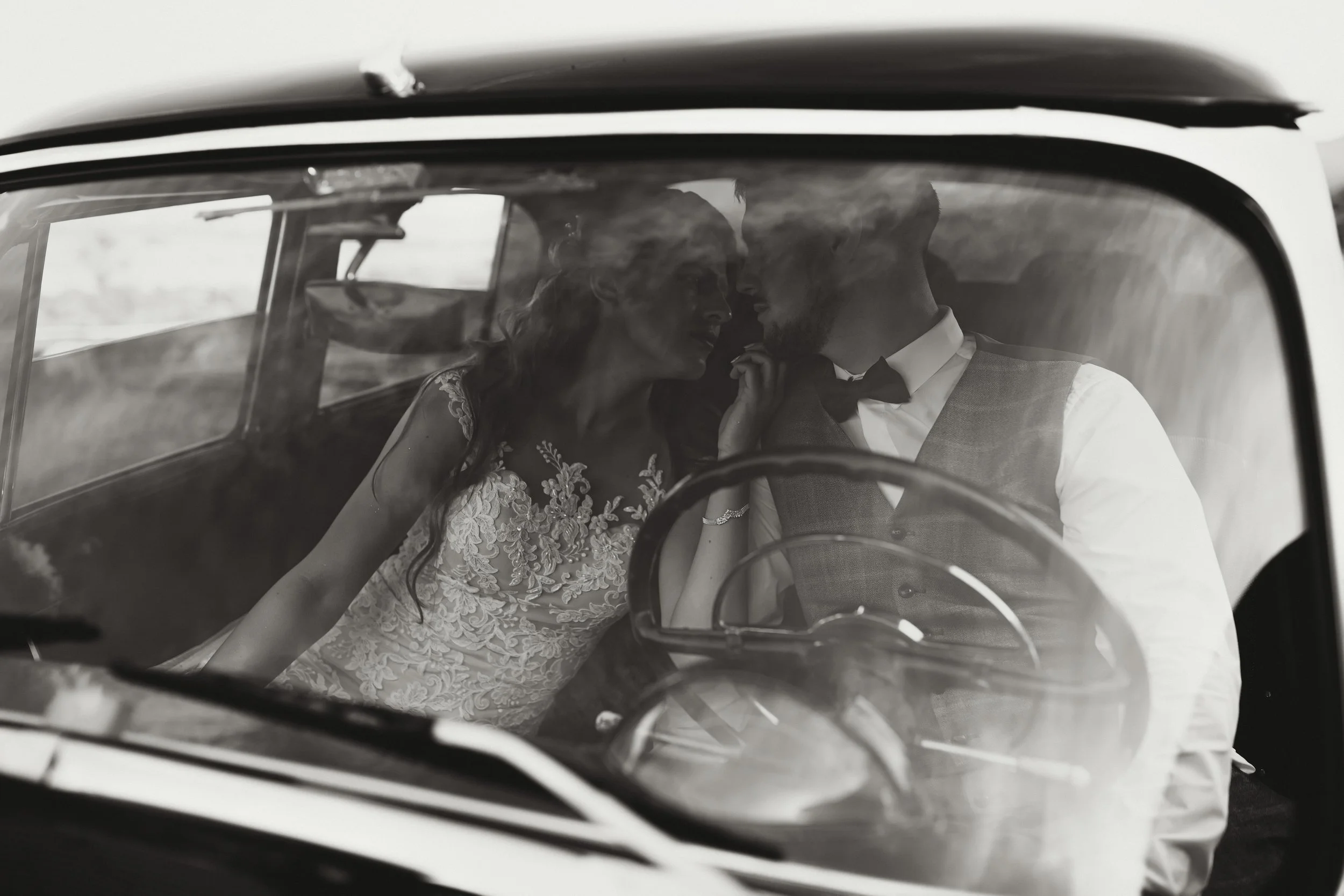 Black and white photo of a couple sitting inside a car, close to each other, with their faces nearly touching. The woman is wearing a floral lace dress, and the man is dressed in a shirt with a vest and bow tie.