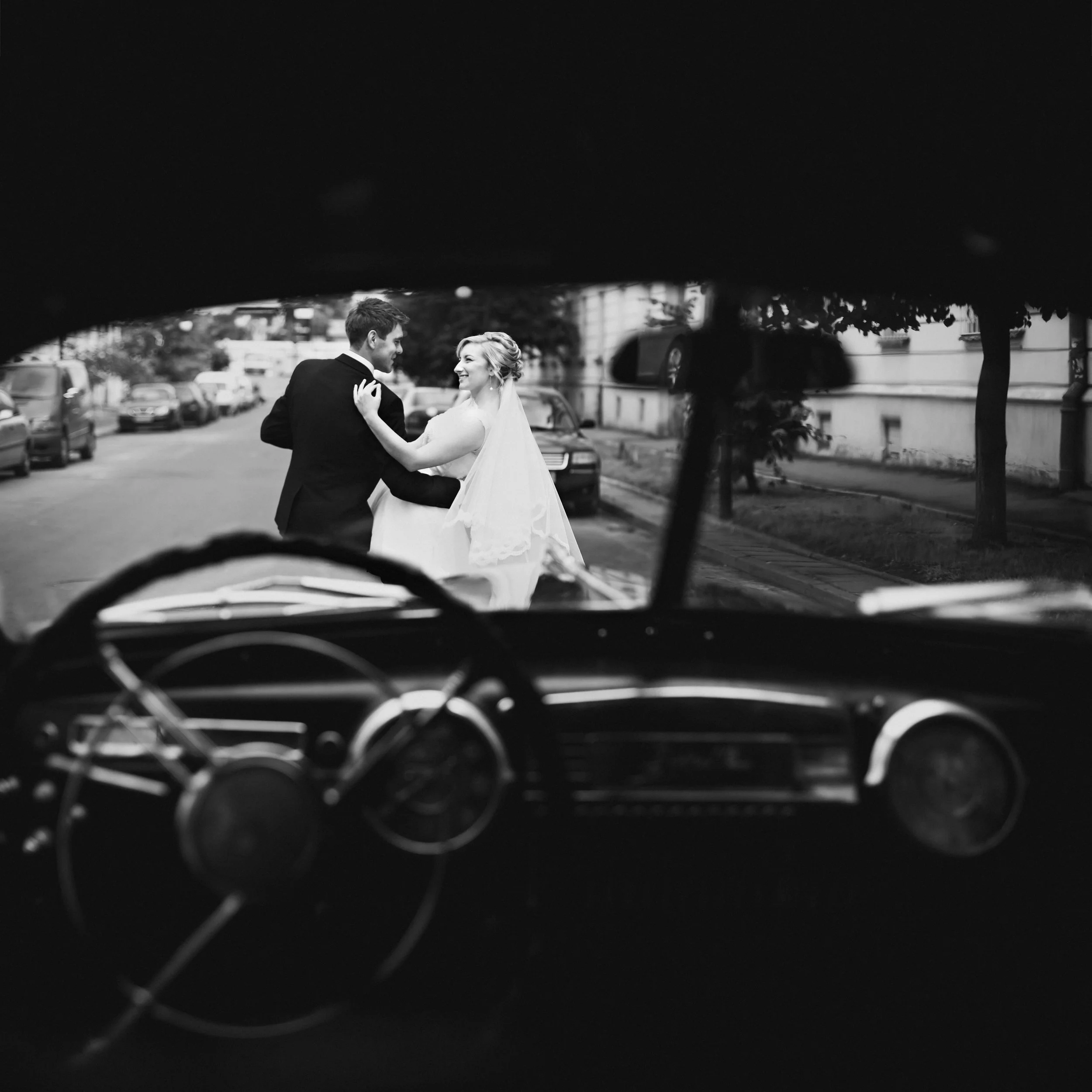 A person in a wedding dress dancing with a man in a tuxedo, viewed through the outline of a car's steering wheel and windshield in black and white.