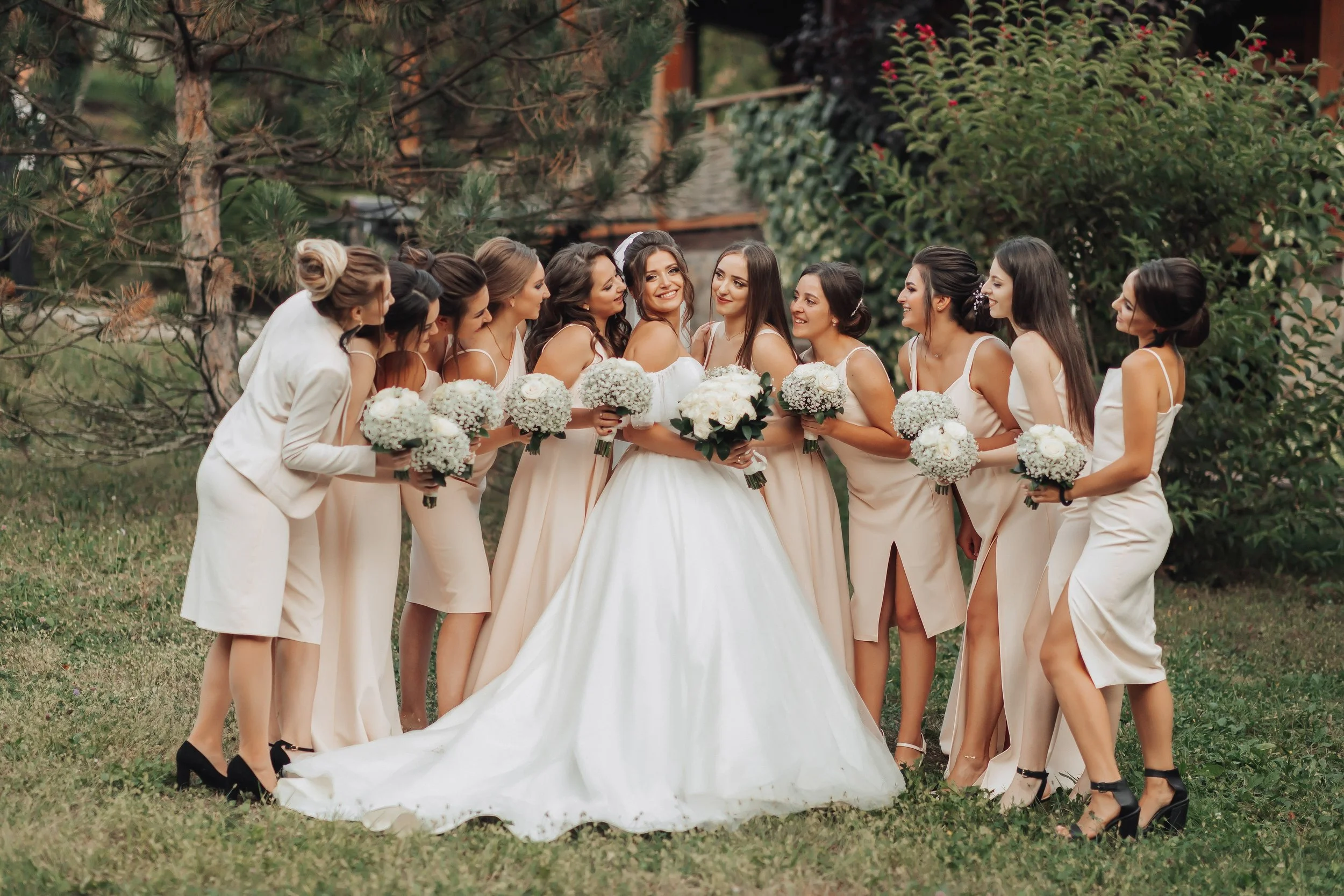 A bride in a white wedding gown surrounded by nine bridesmaids in matching beige dresses holding bouquets, outdoors in a garden area with trees and shrubbery.