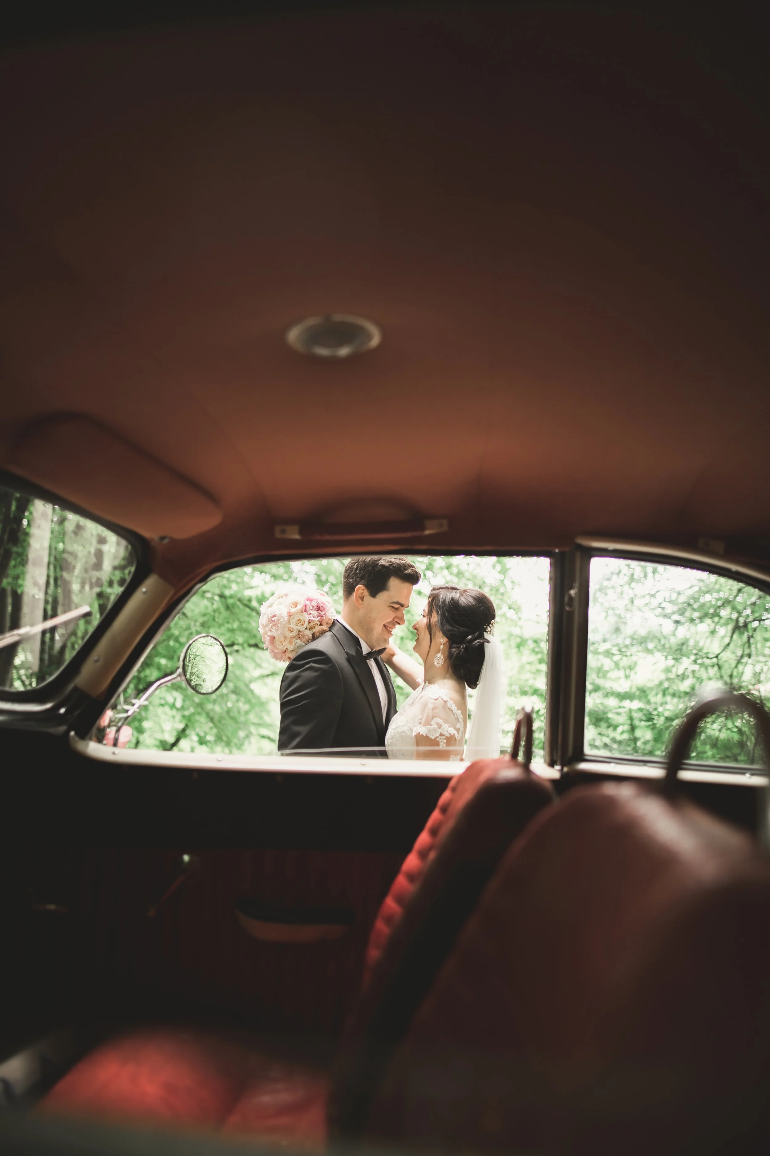A bride and groom smiling at each other, visible through the window of a vintage car, with lush green trees in the background.