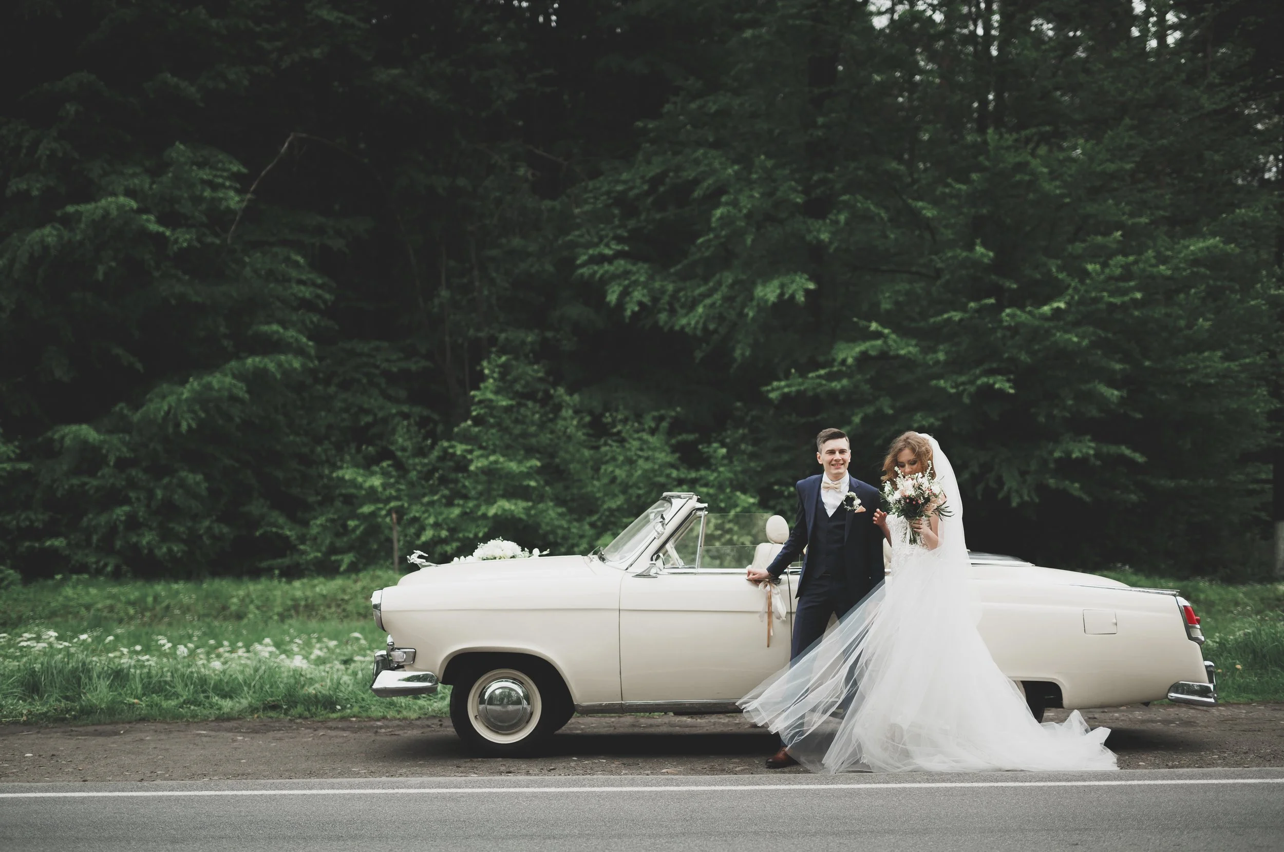 A bride and groom standing beside a white vintage convertible car decorated with flowers on a grassy roadside, with a dark green forest in the background.